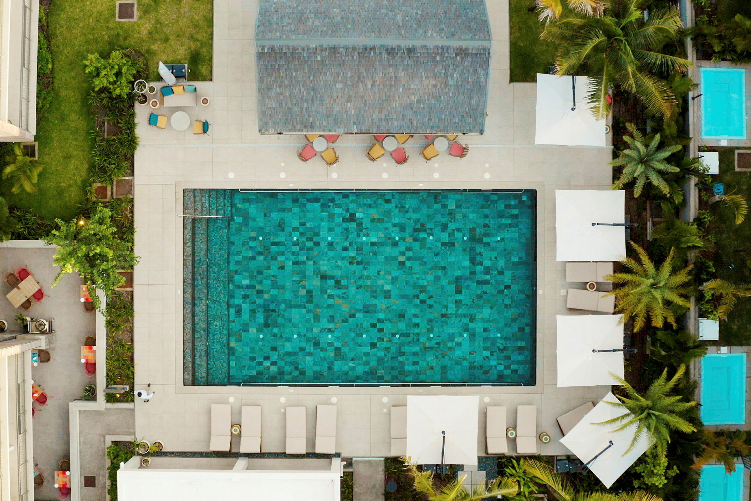 Luftfoto af en rektangulær pool omgivet af liggestole, parasoller og grønne planter. (AI-genereret tekst)