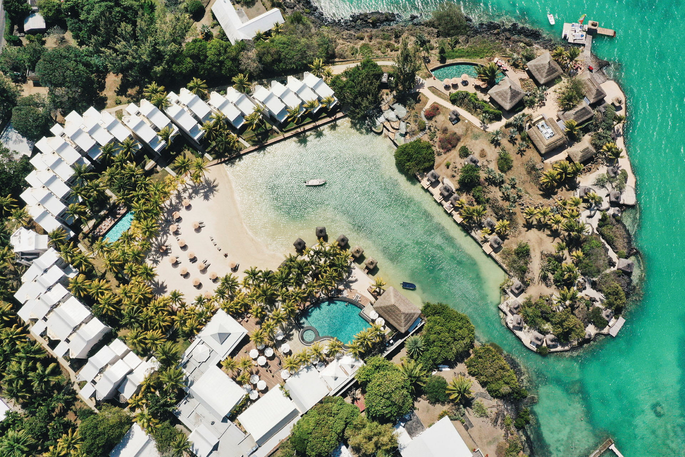 Luftfoto af et hotelområde med pool, en sandstrand og beplantning ved vandet. (AI-genereret tekst)