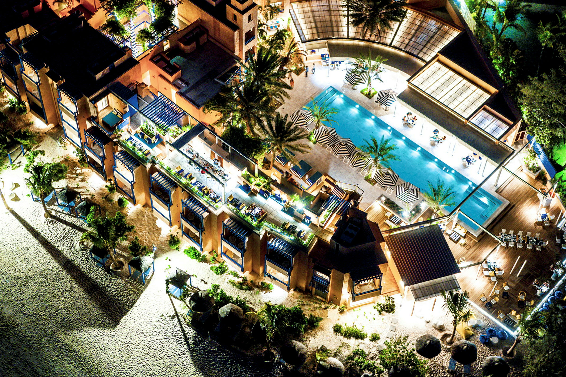 Fugleperspektiv over en resort med pool, liggestole, parasoller og omkringliggende bygninger. (AI-genereret tekst)