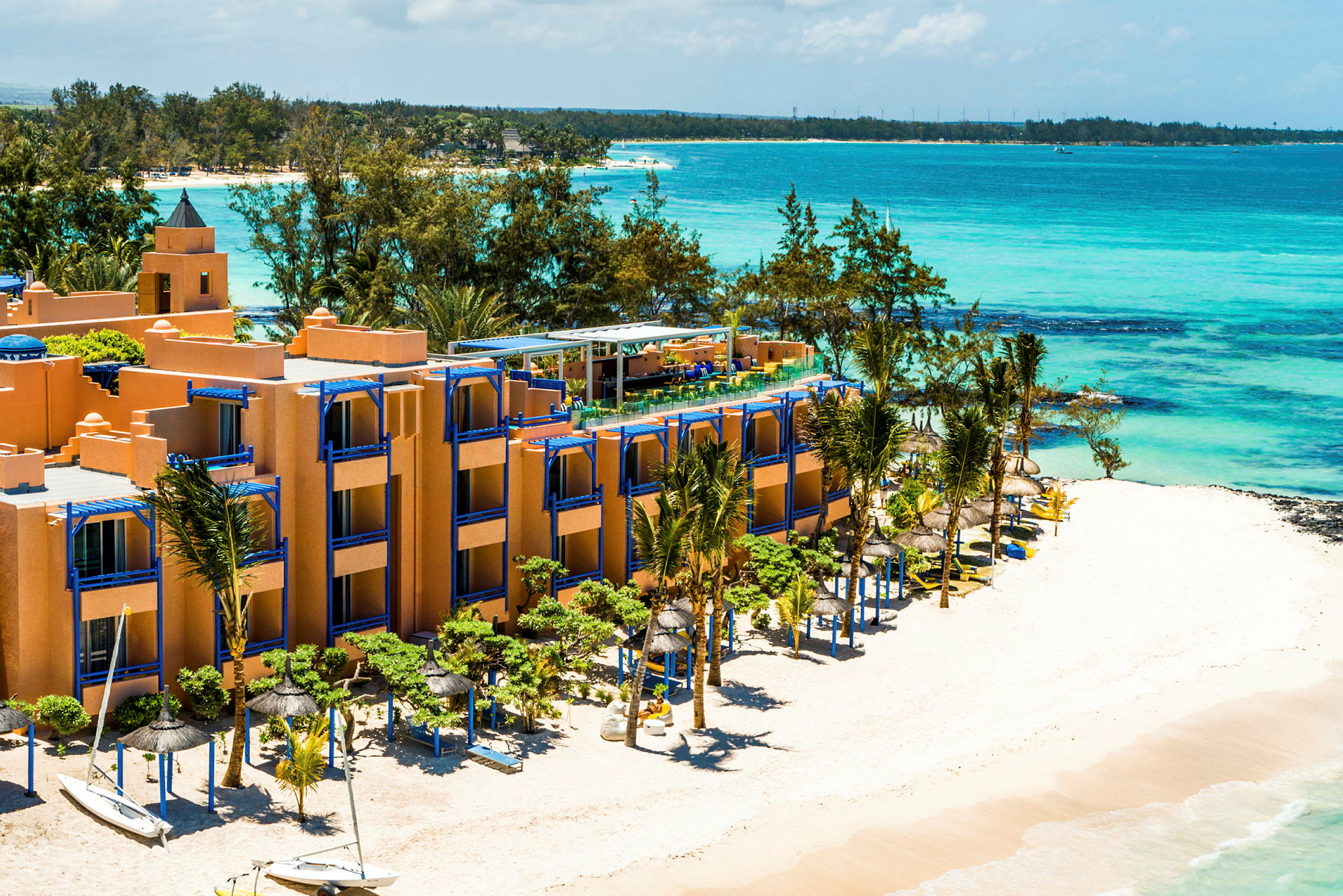 Hotelbygning ved en hvid sandstrand med blå parasoller og havet i baggrunden. (AI-genereret tekst)