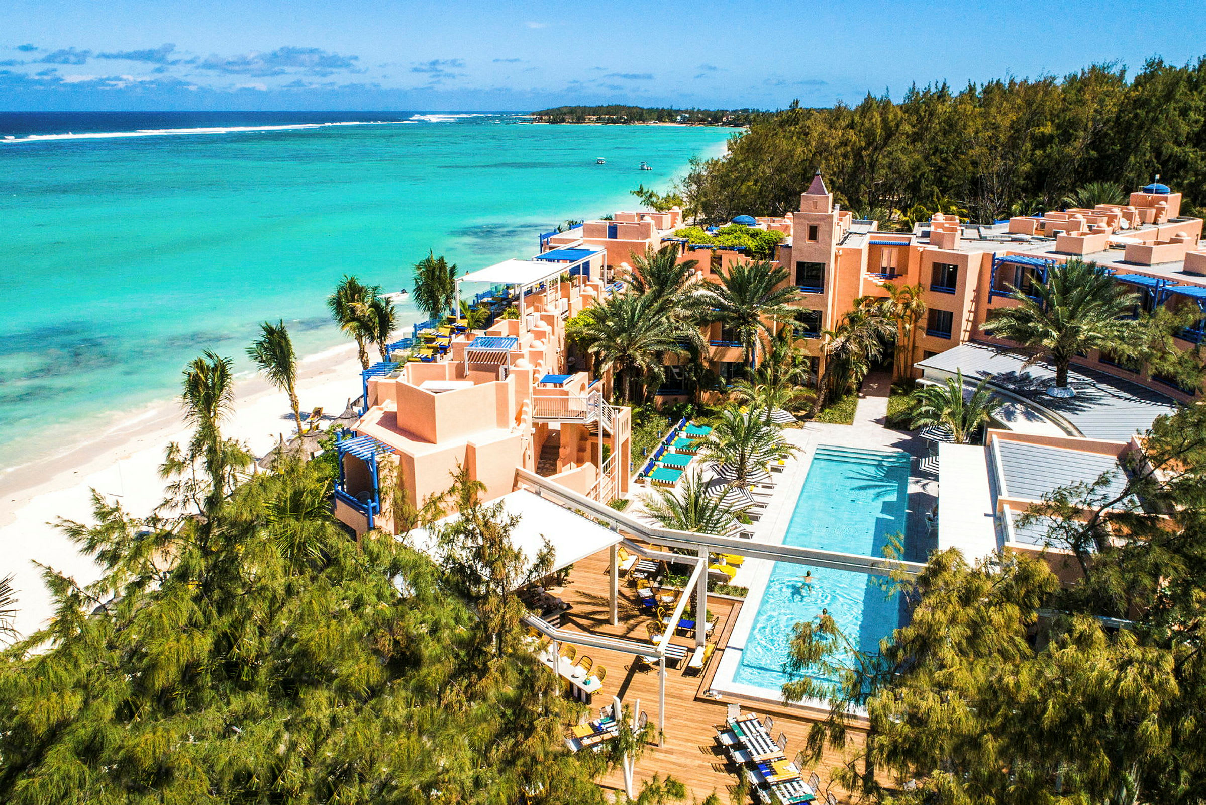 Luftfoto af et hotelkompleks ved en tropisk strand med pool og omkringliggende vegetation. (AI-genereret tekst)
