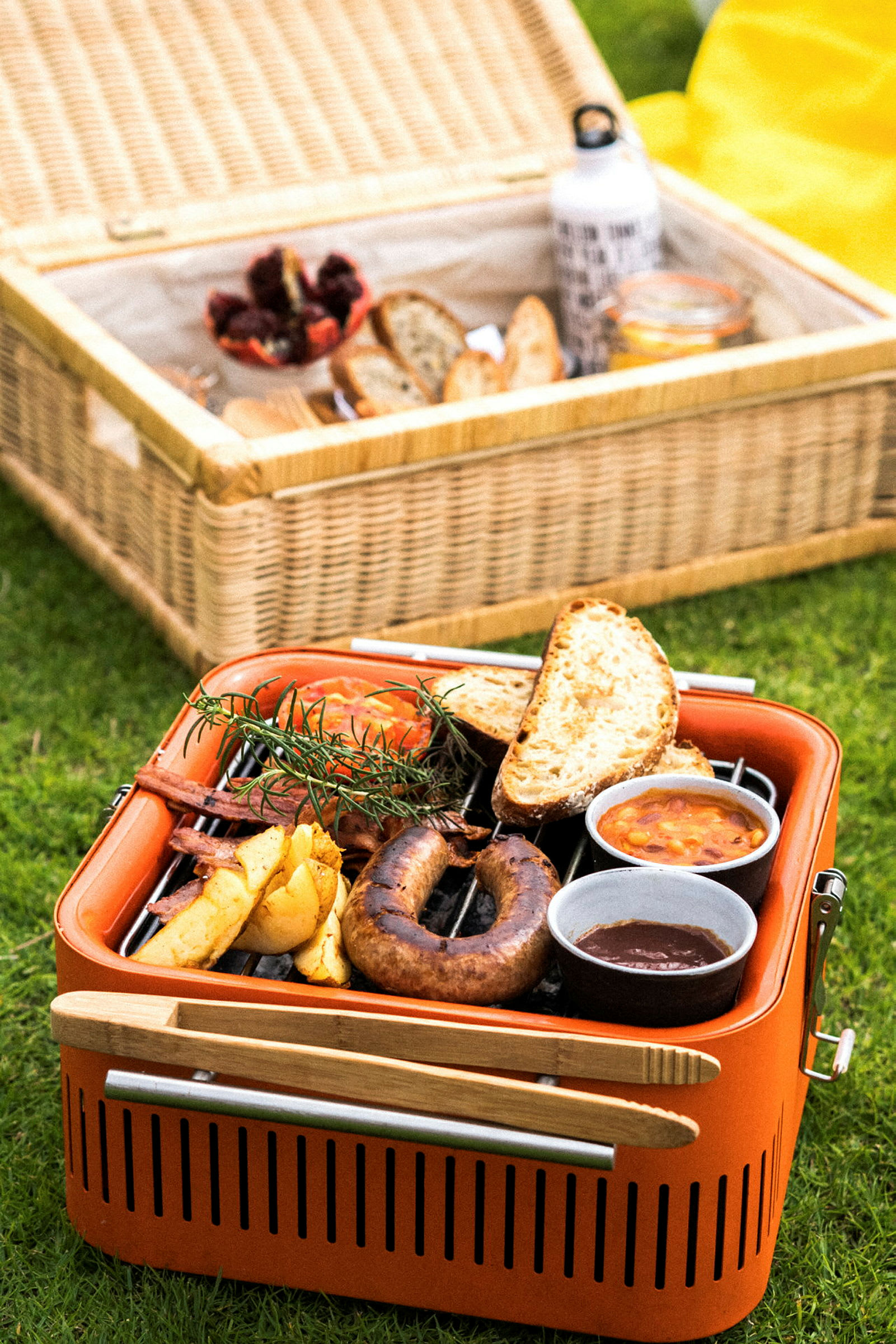 En orange grill med mad, inklusive pølser og brød, står på græsset. I baggrunden er en kurv med picnictilbehør. (AI-genereret tekst)