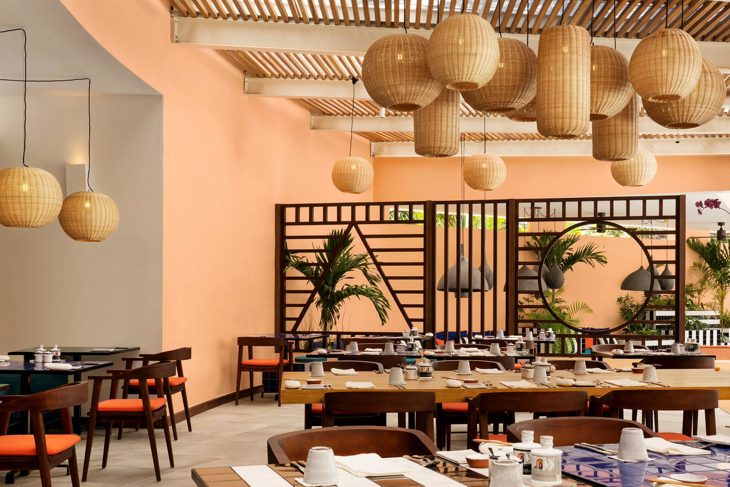 En indendørs restaurant med dækkede borde og hængende kurvelamper. Baggrunden viser orange vægge og grønne planter. (AI-genereret tekst)