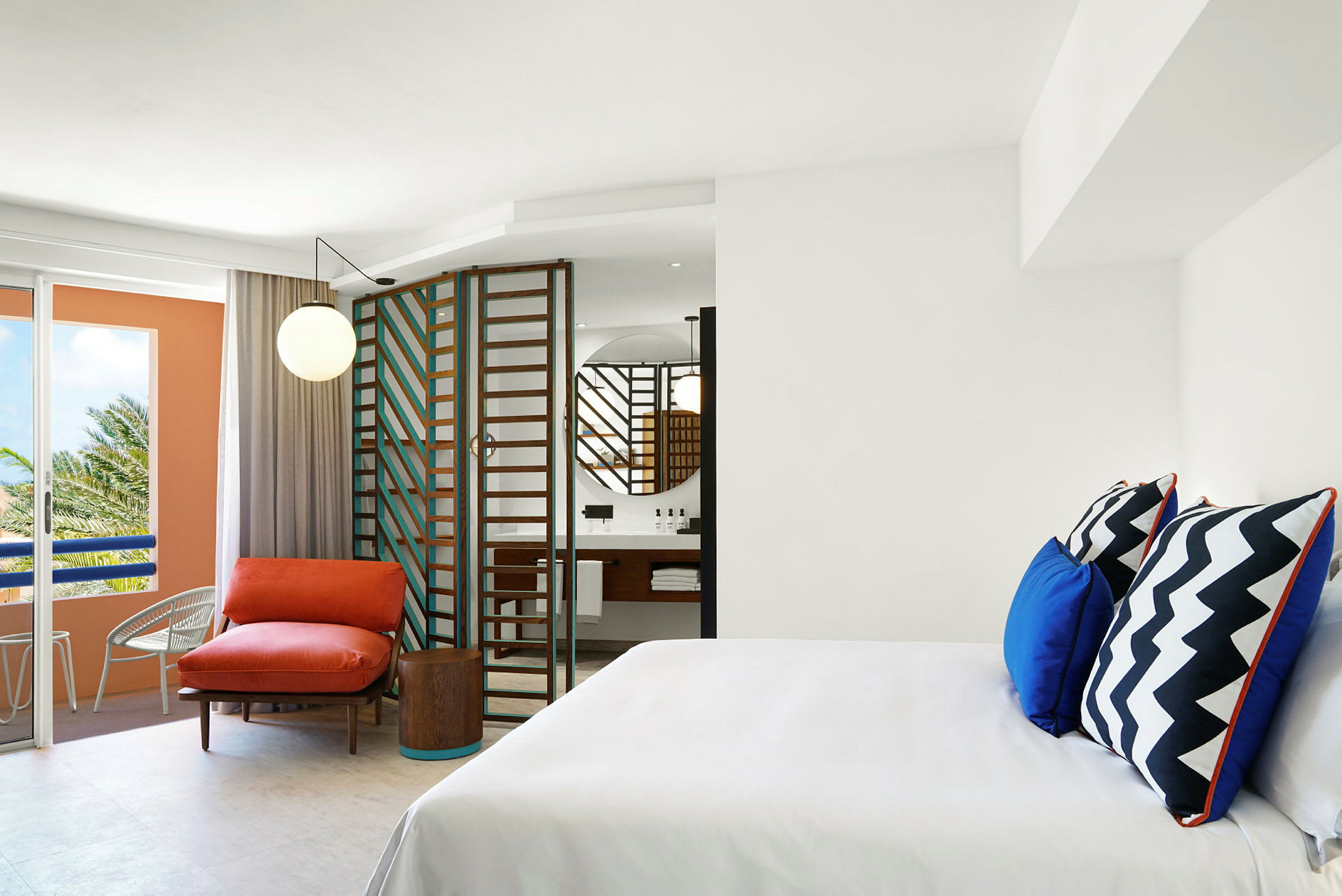 Et moderne hotelværelse med hvide vægge, en farverig lænestol og puder samt et badeværelse med spejl og en glasdør. (AI-genereret tekst)