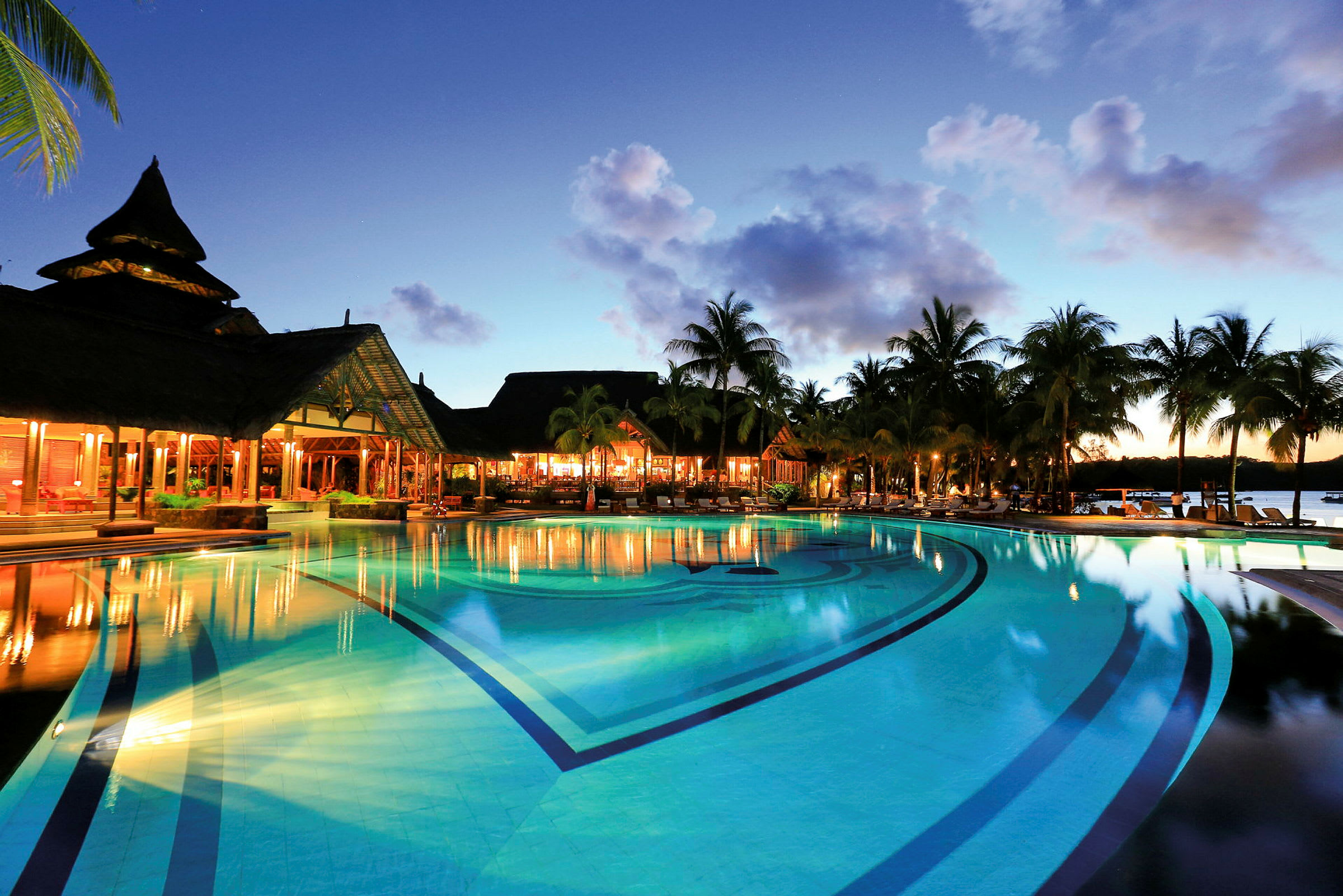 Pool ved en resort ved solnedgang med palmer og oplyste bygninger. (AI-genereret tekst)