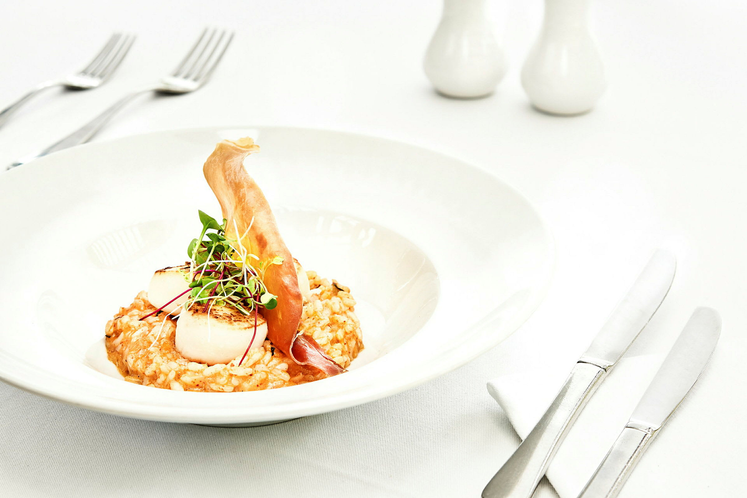Tallerken med risotto, grillede kammuslinger og en dekoration af mikrogrønt samt tynde skiver ingredienser. (AI-genereret tekst)