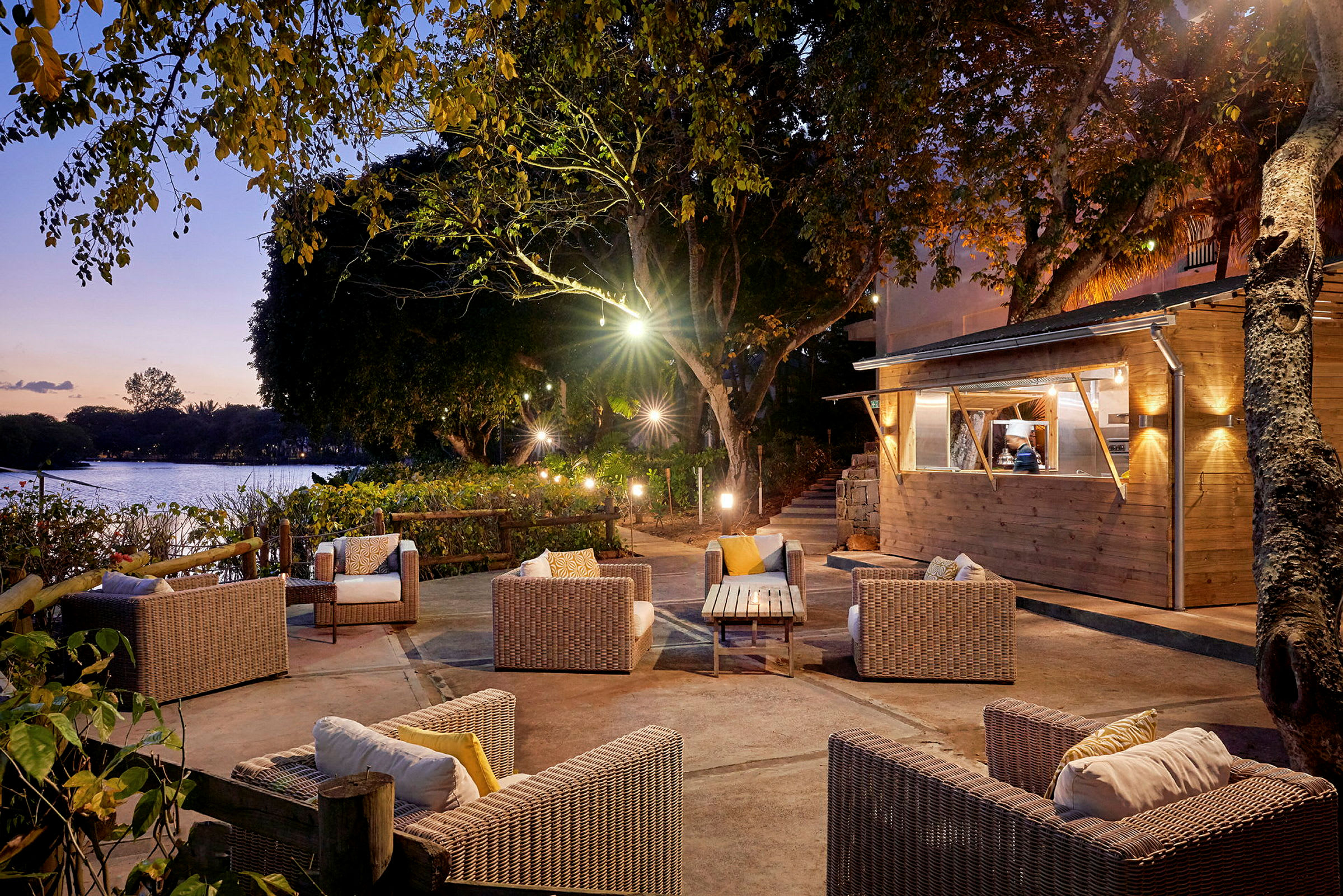 En aftenbelyst terrasse med sofaarrangement og et træhus ved siden af en sø. (AI-genereret tekst)