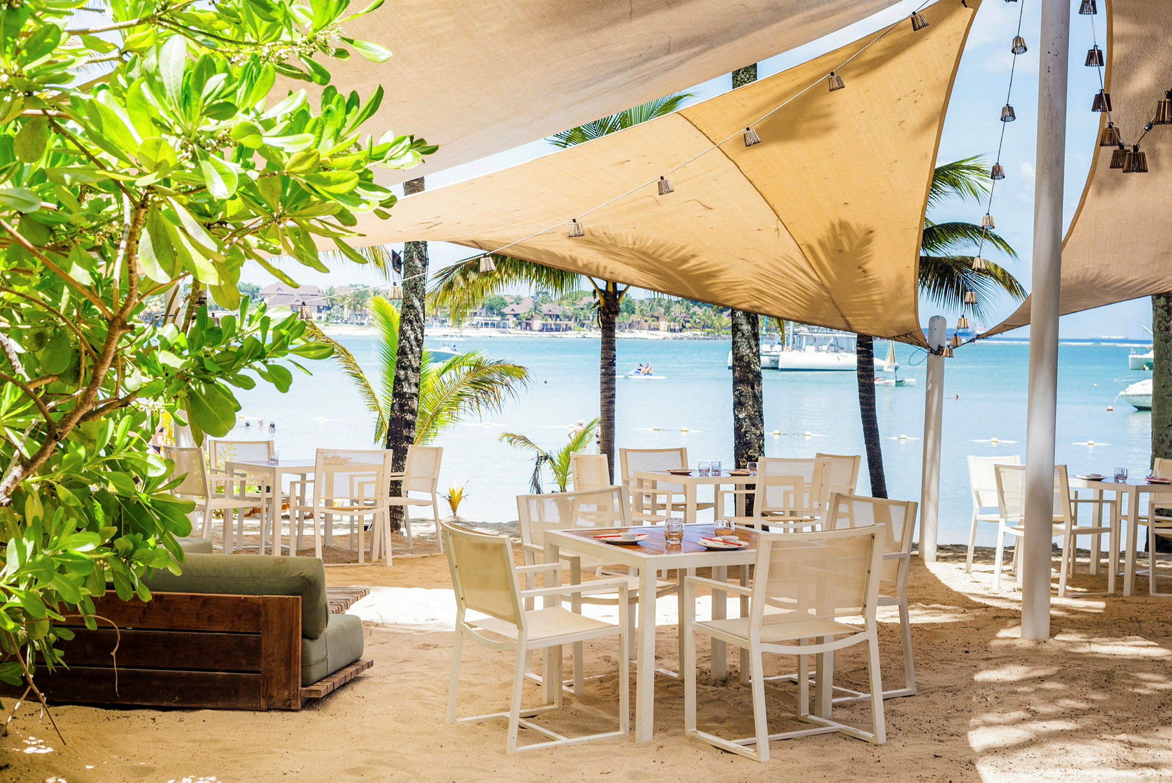 Udendørs restaurant med borde og stole på sandstrand, omgivet af grønt og palmer med udsigt over havet. (AI-genereret tekst)