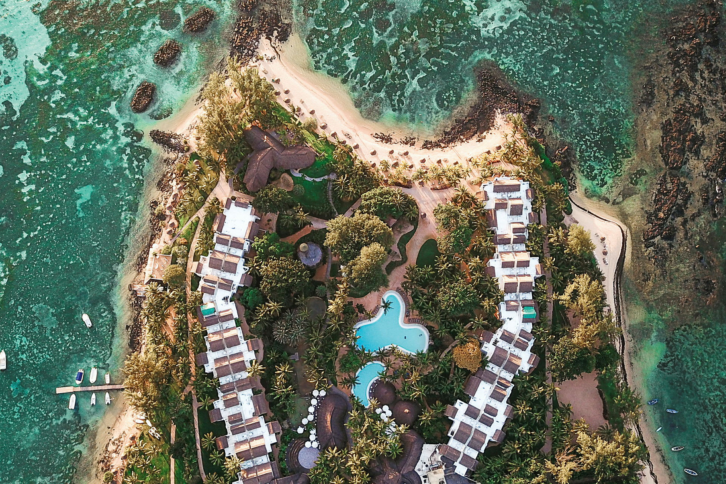 Luftfoto af en ferieby med pool og omkringliggende grønne områder ved kysten. (AI-genereret tekst)