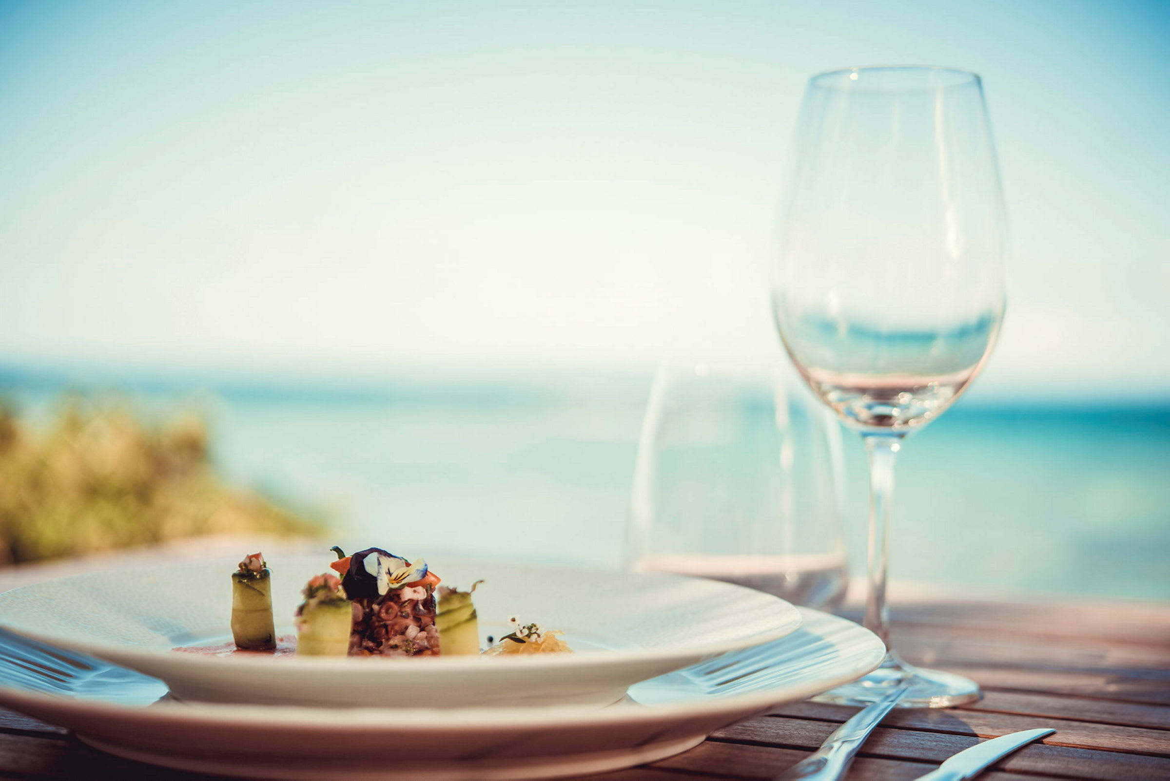 En tallerken med en elegant ret og et glas på et bord med havudsigt. (AI-genereret tekst)