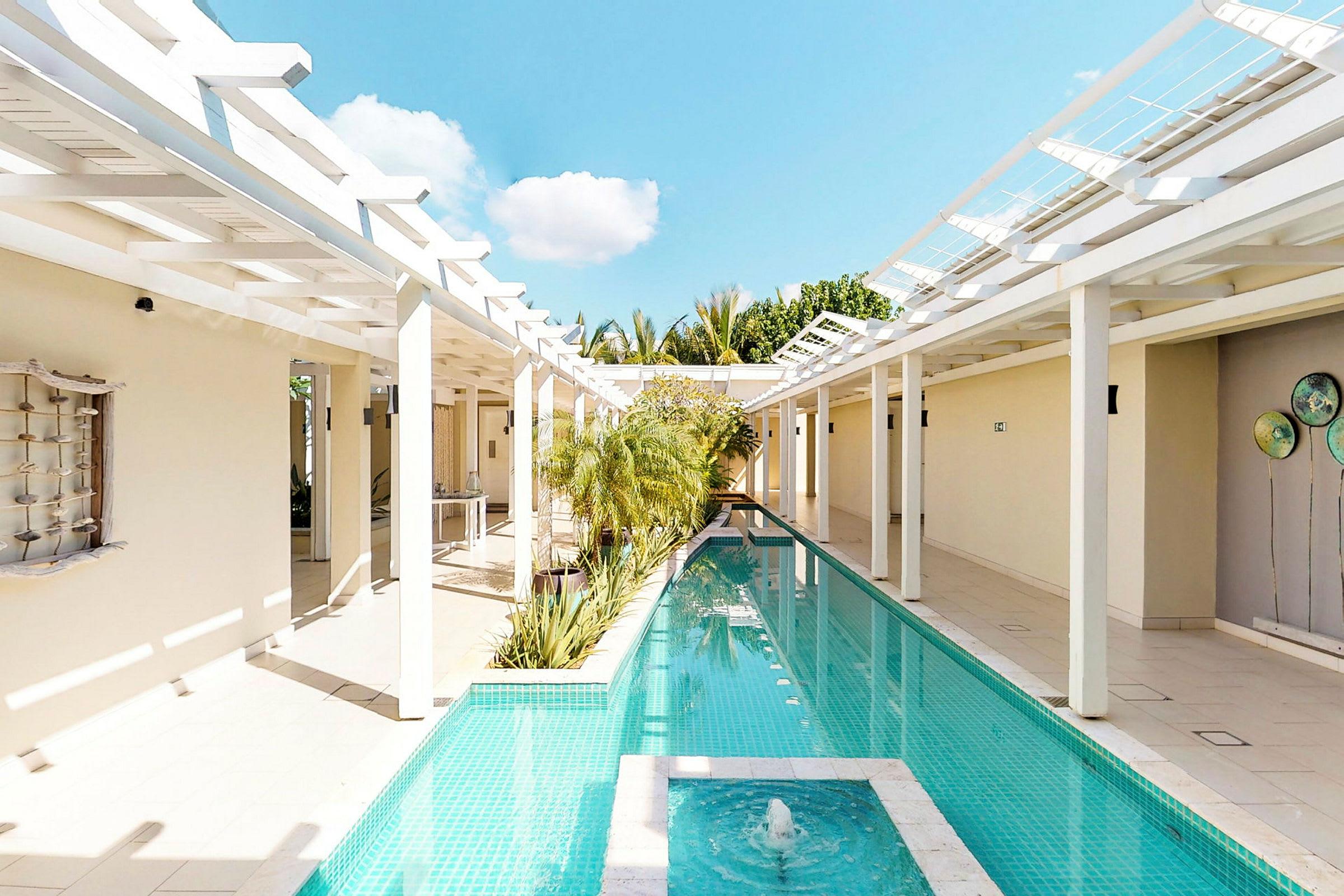En lang, smal pool omgivet af hvide søjler og pergola med en fontæne i forgrunden. (AI-genereret tekst)