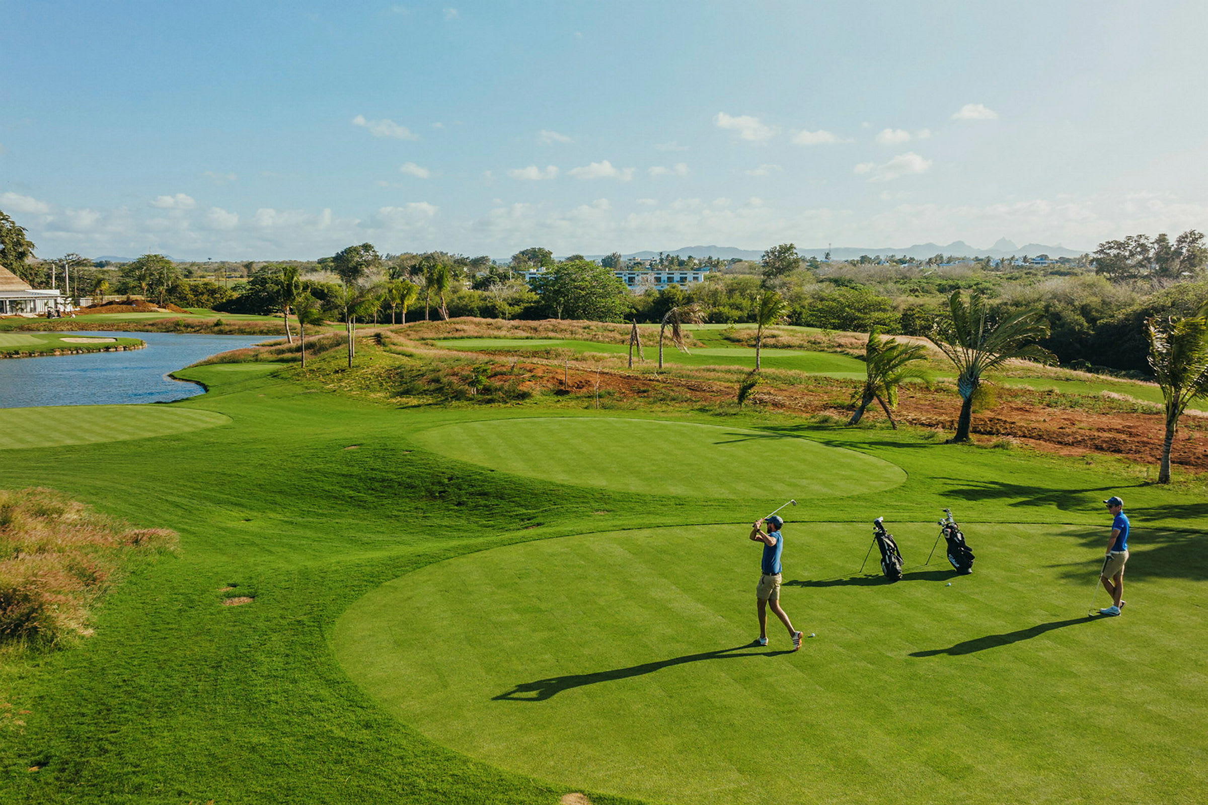 To personer spiller golf på en grøn golfbane med en lille sø og træer i baggrunden. (AI-genereret tekst)