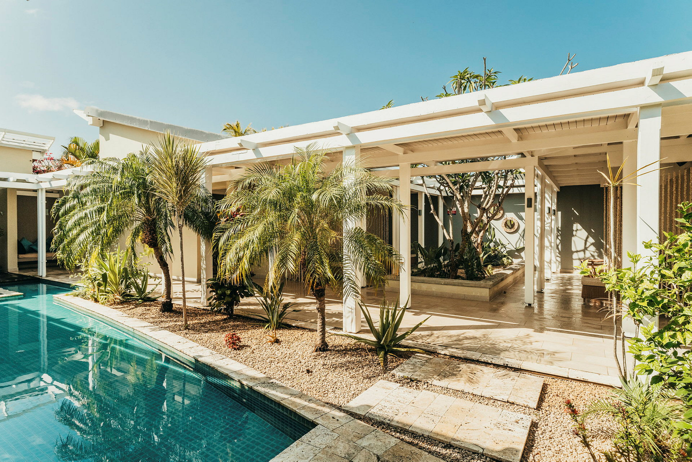 Et udendørsmiljø med pool, palmer og en bygning med hvid pergola. (AI-genereret tekst)
