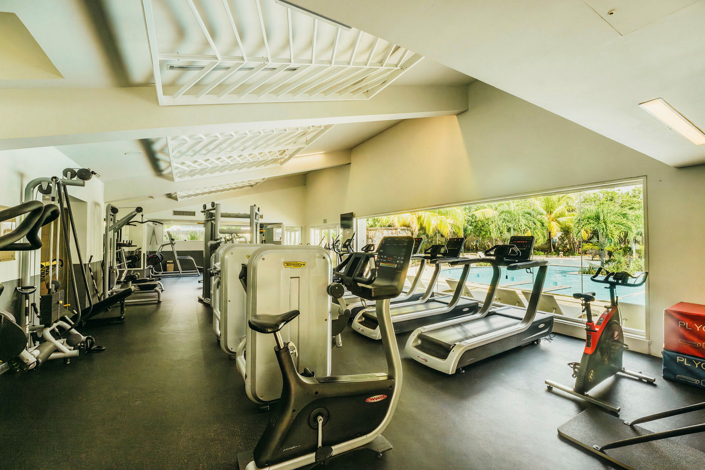 Træningscenter med fitnessmaskiner og udsigt til en udendørs pool. (AI-genereret tekst)