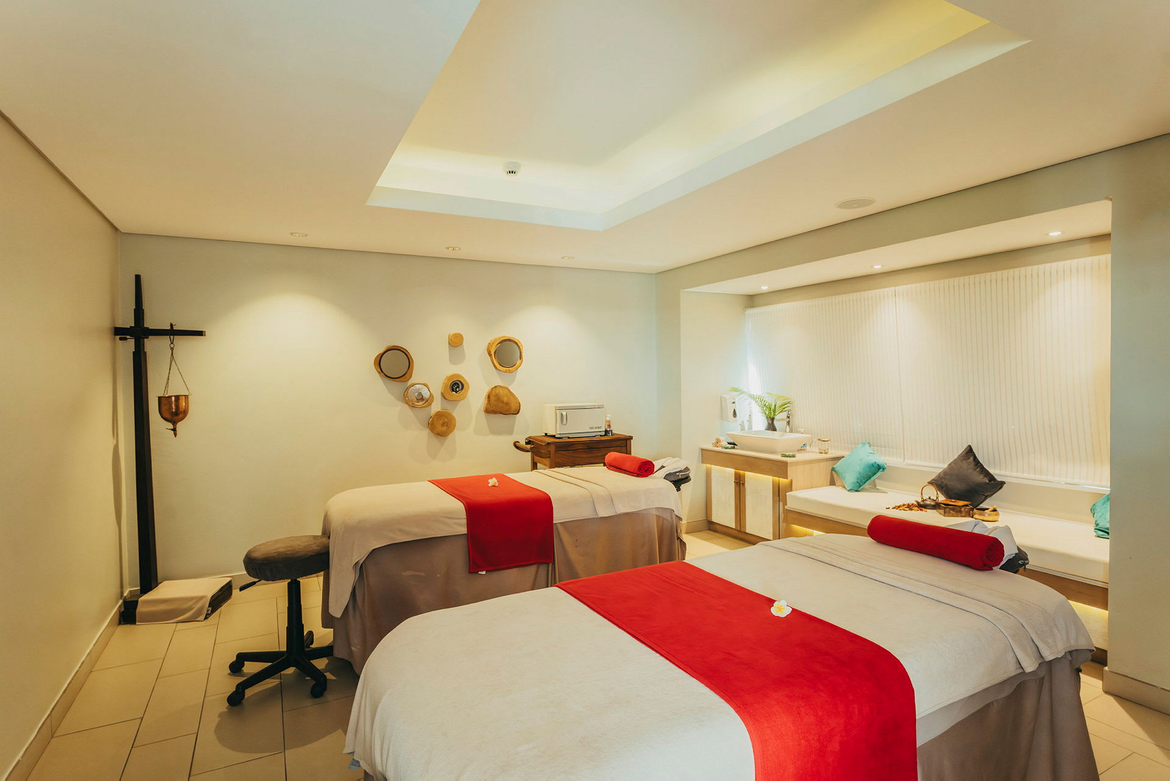 Et spa-rum med to massageborde dækket med beige lagner og røde detaljer, indrettet med dekorative elementer og moderne belysning. (AI-genereret tekst)