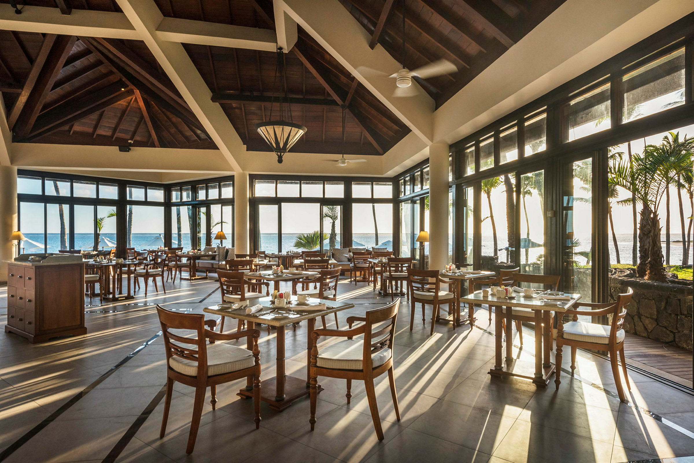 Et åbent restaurantområde med træborde og stole, store vinduer og udsigt over havet og palmer. (AI-genereret tekst)
