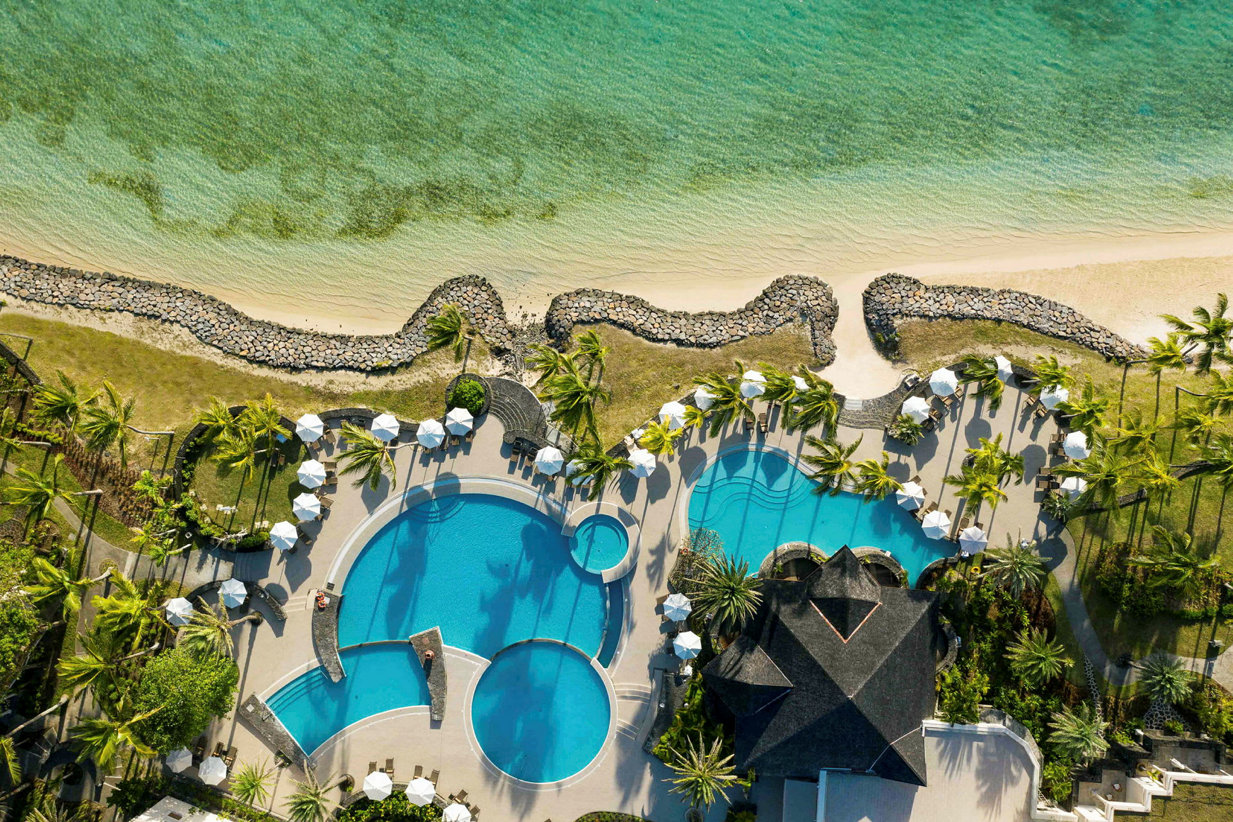 Luftfoto af et hotelområde med swimmingpools, liggestole og parasoller, omgivet af grønne områder tæt på en kystlinje. (AI-genereret tekst)