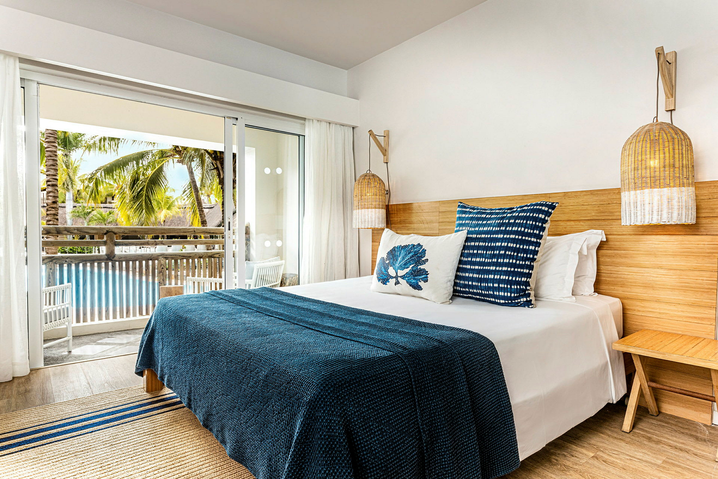 Et hotelværelse med en seng med blåt sengetæppe og dekorative puder. Værelset har en balkon med udsigt over en pool og palmer. (AI-genereret tekst)