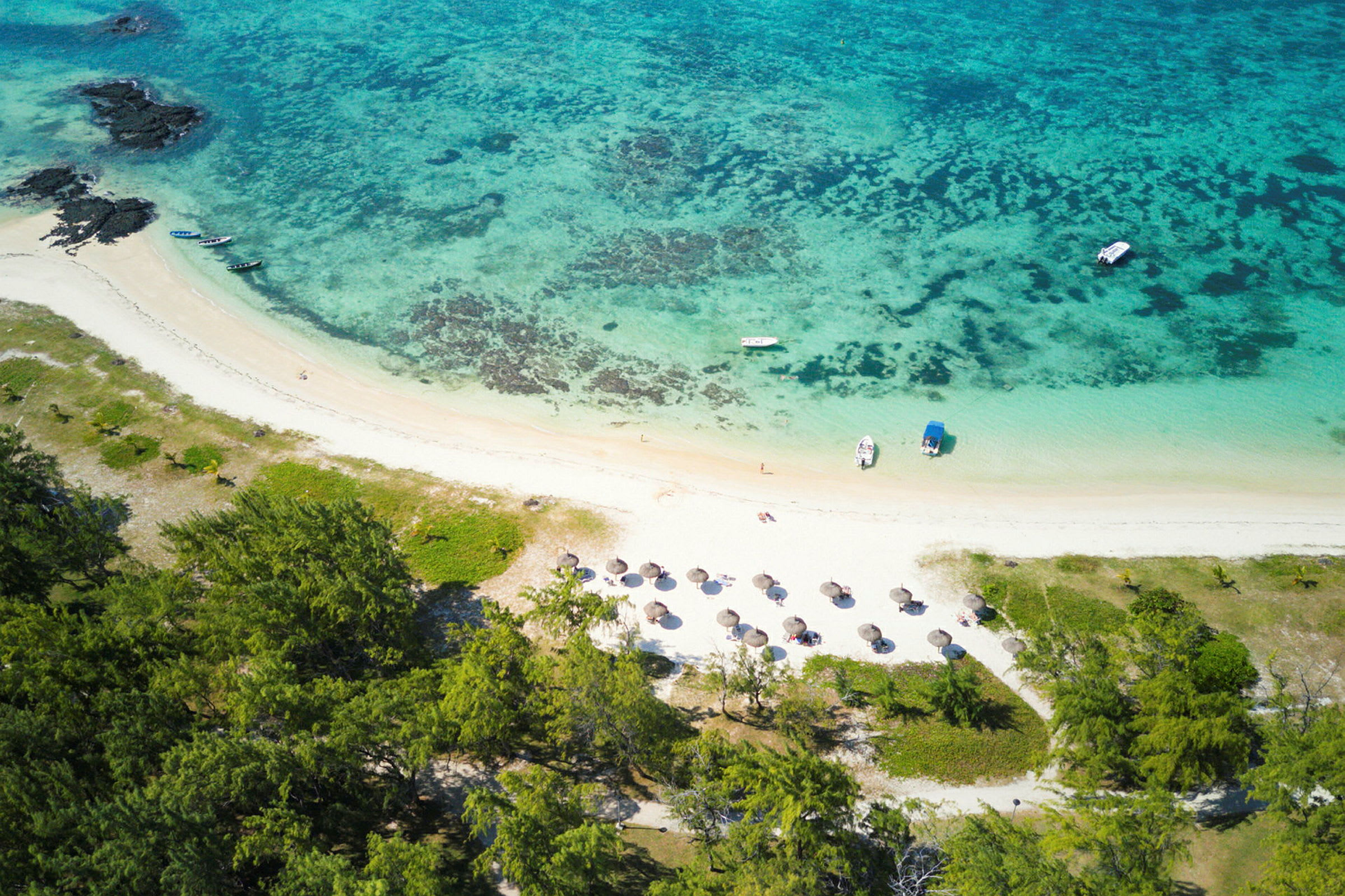 Luftfoto af en tropisk strand med parasoller, både og omkringliggende grønne områder. (AI-genereret tekst)