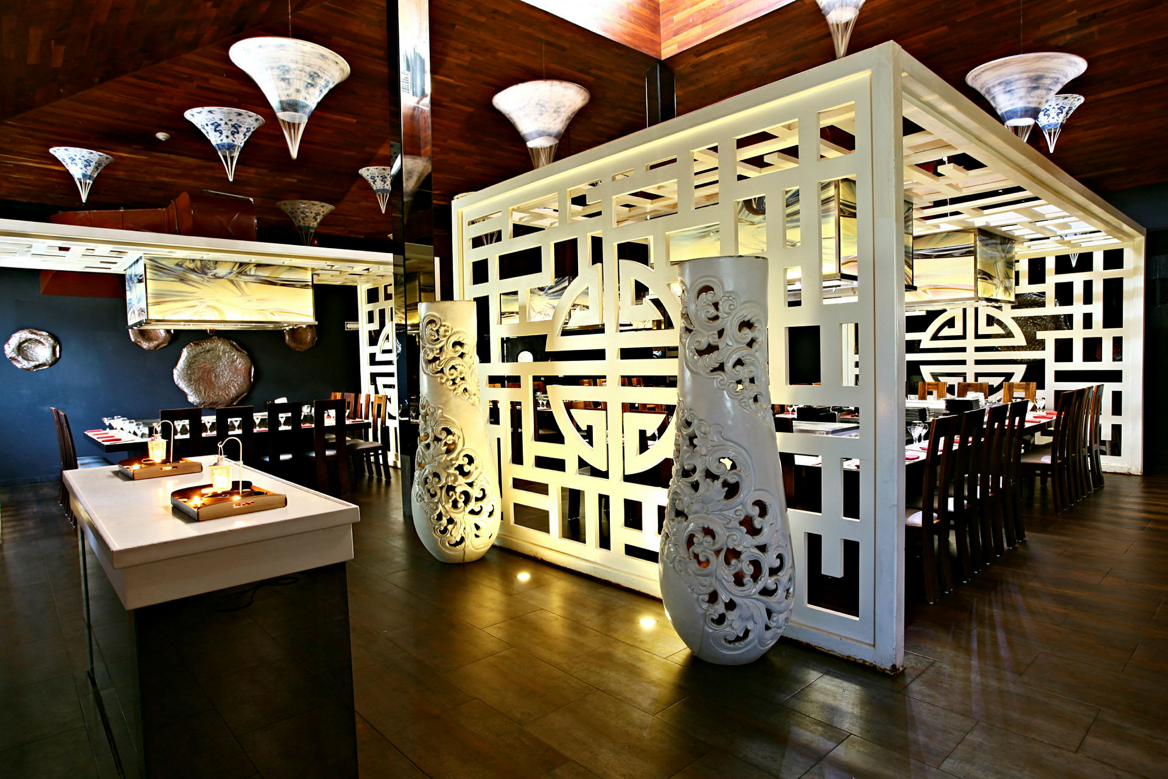 En indendørs restaurant med et elegant indrettet område med træborde og stole. Rummet er dekoreret med hvide ornamenterede skillevægge og hængende lamper. (AI-genereret tekst)