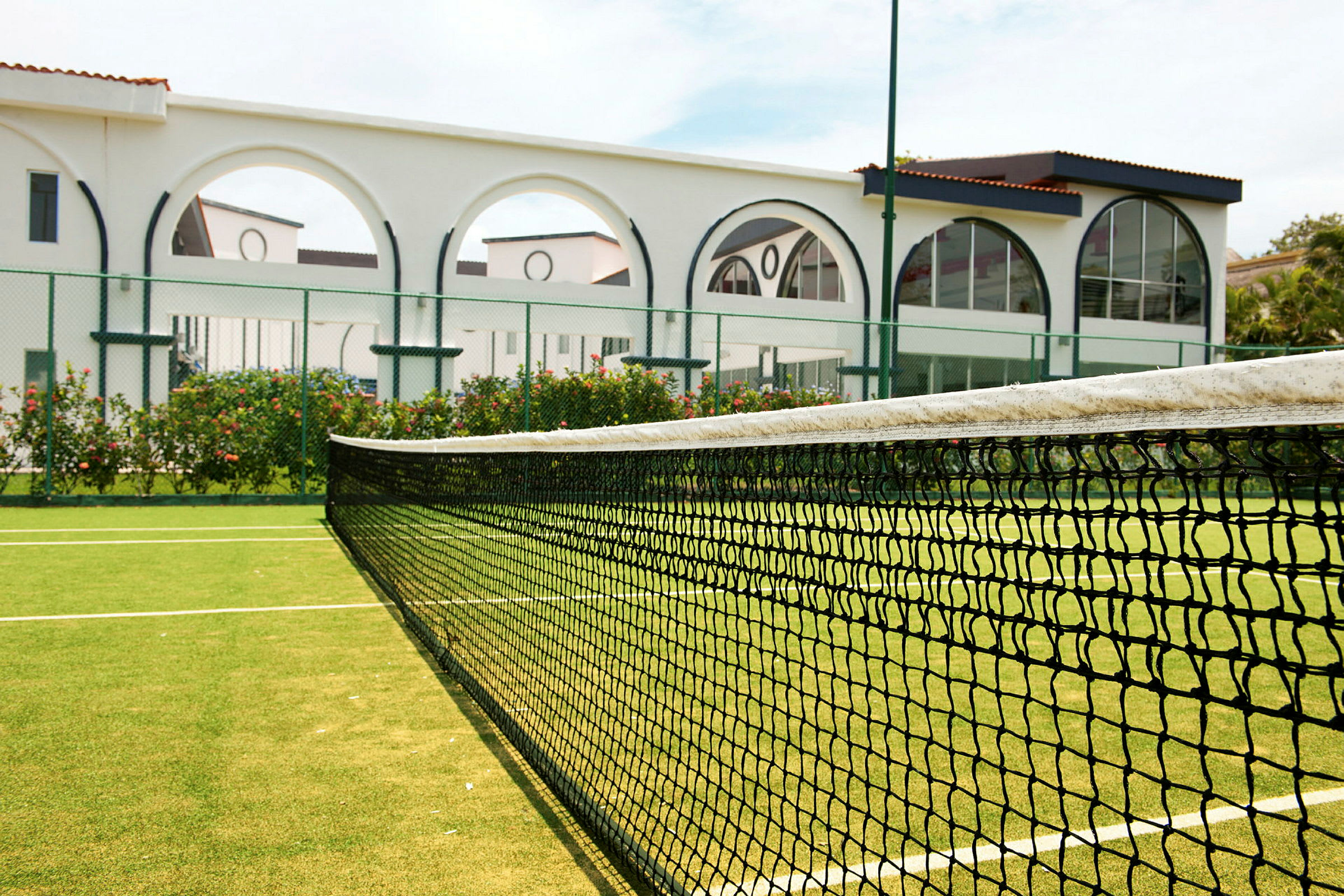 En tennisbane med en grøn overflade og et net i forgrunden. I baggrunden ses en hvid bygning med buer. (AI-genereret tekst)