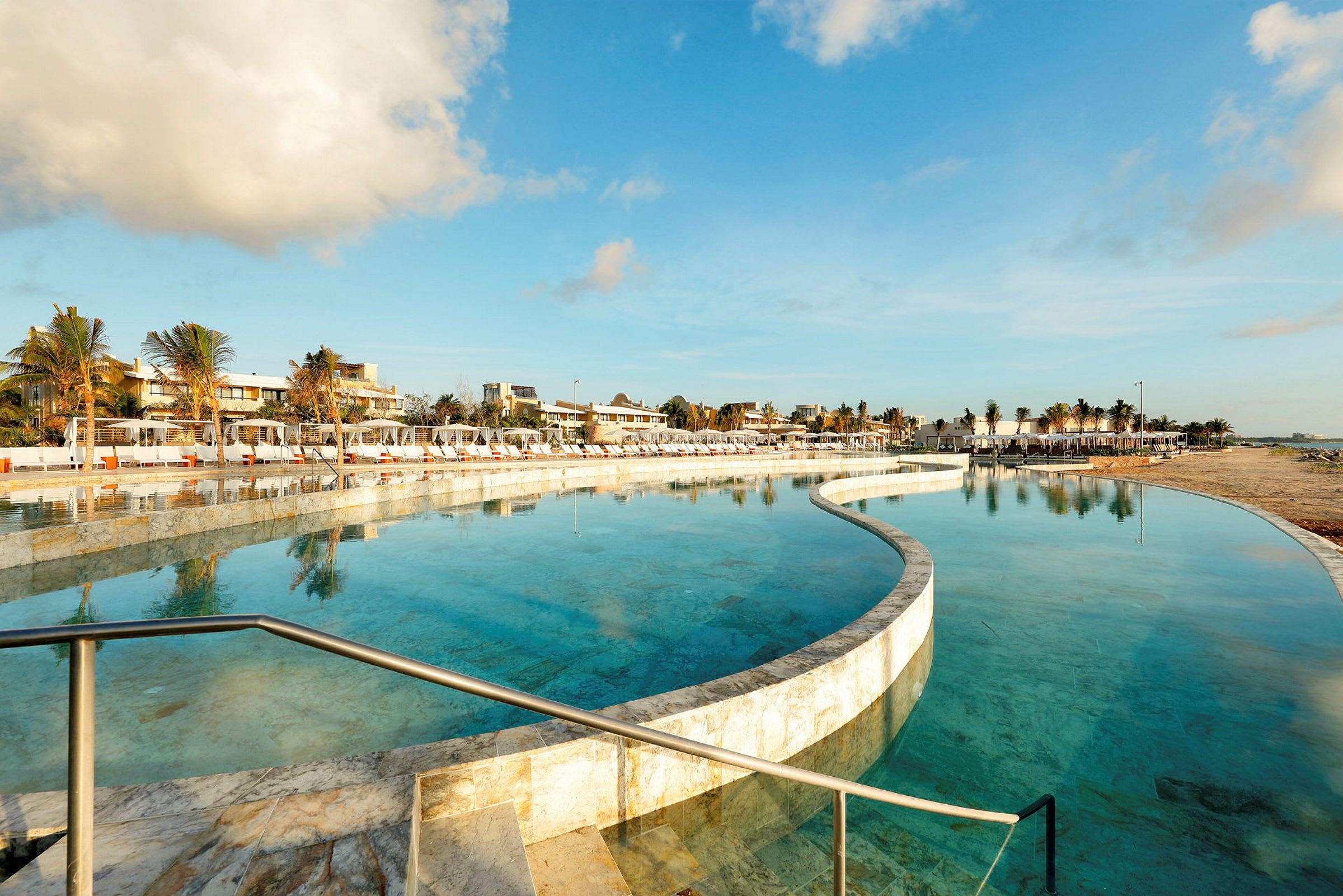 En strandnær pool omgivet af liggestole og palmer med klar himmel over. (AI-genereret tekst)
