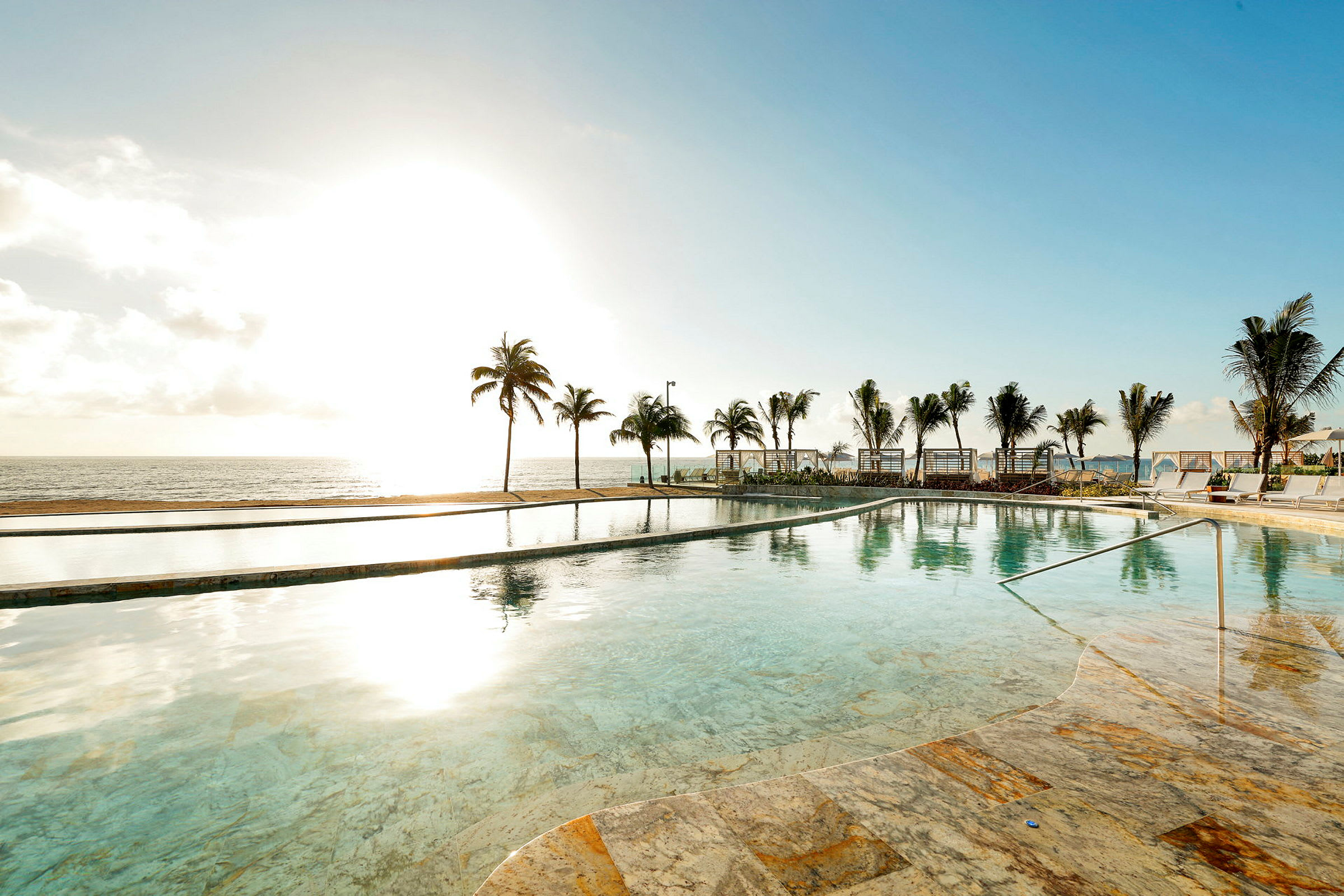 En infinitypool med klart blåt vand ligger foran en strand omgivet af palmer og liggestole. (AI-genereret tekst)