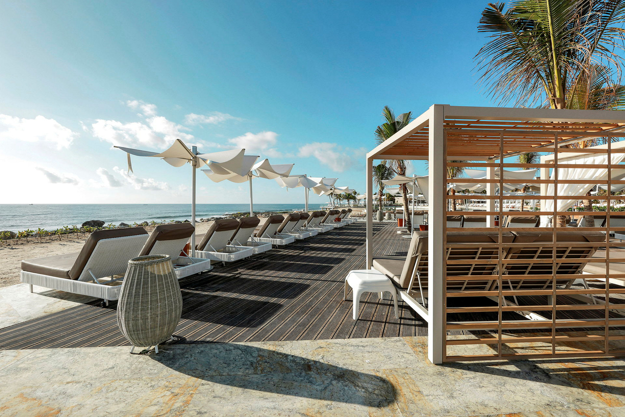 En strand med liggestole, parasoller og en pergola i forgrunden under en blå himmel. (AI-genereret tekst)