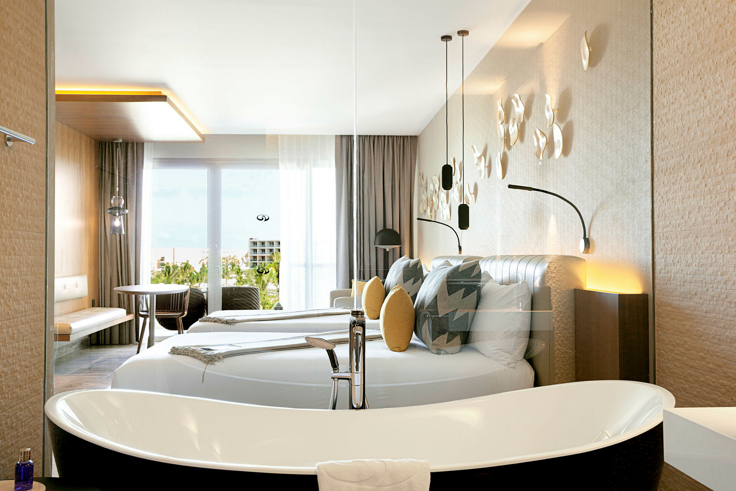 Et moderne hotelværelse med en stor dobbeltseng, dekorative puder og vægdekorationer. Der er et fritstående badekar og møbler som bord og stole nær en glasdør med udsigt. (AI-genereret tekst)
