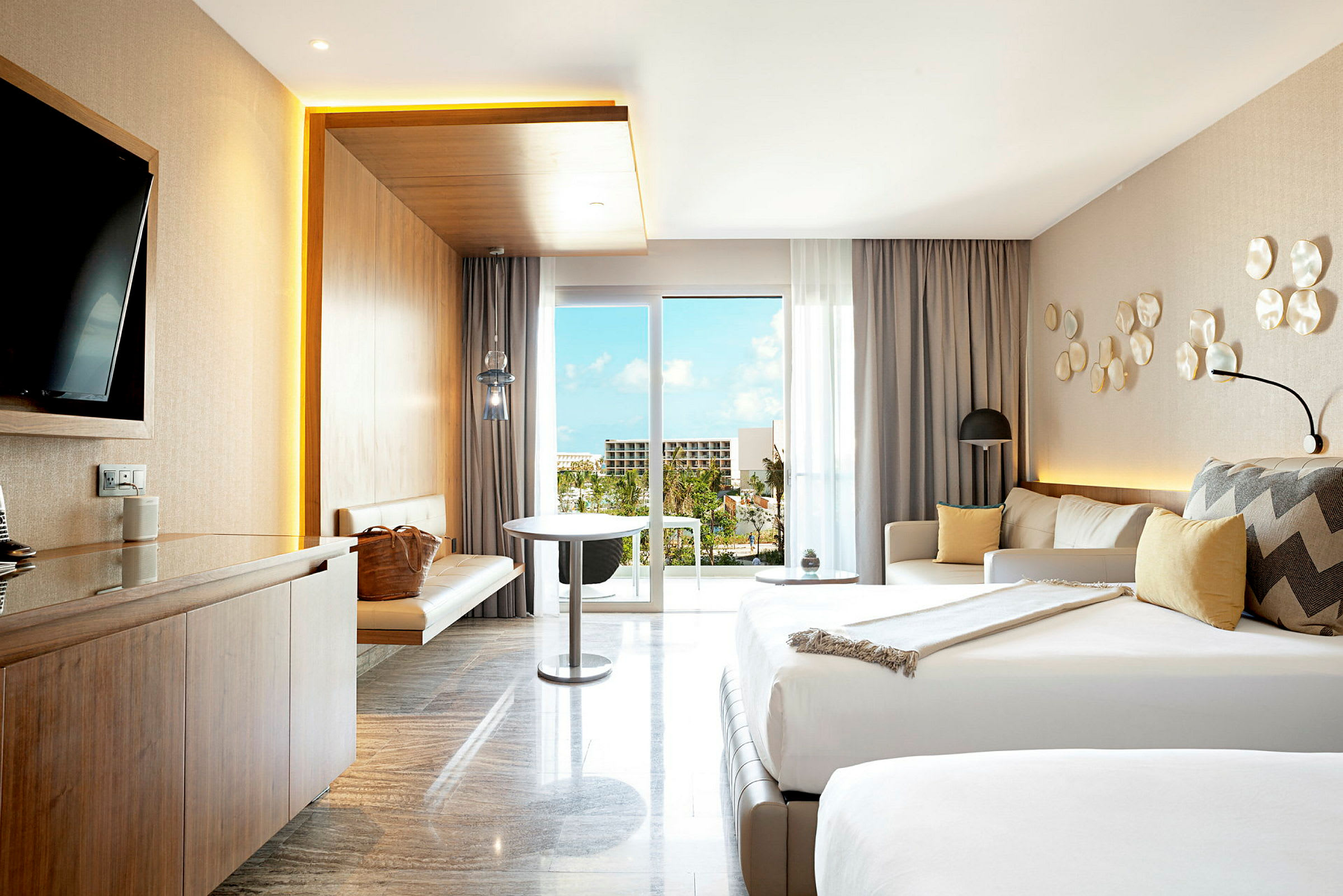 Et moderne hotelværelse med dobbeltseng, sofagruppe, bord og TV. Lyse farver og en balkon med udsigt over natur og bygninger. (AI-genereret tekst)