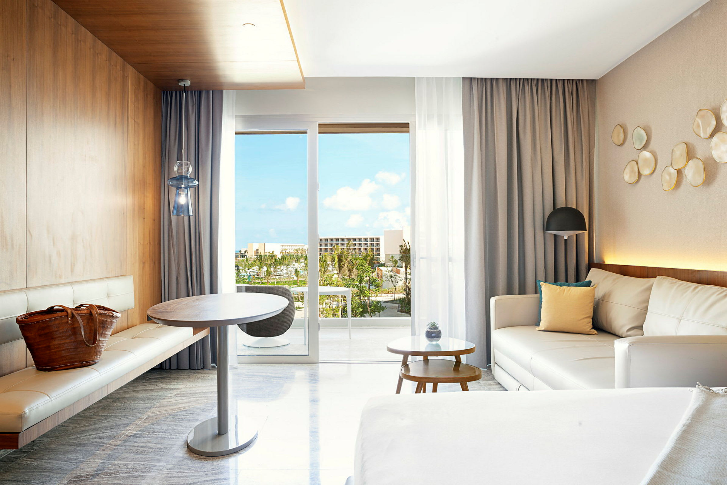 Et moderne hotelværelse med hvide møbler og træpaneler. Udsigt fra panoramavinduerne på altanen med tropiske planter og blå himmel synlig. (AI-genereret tekst)