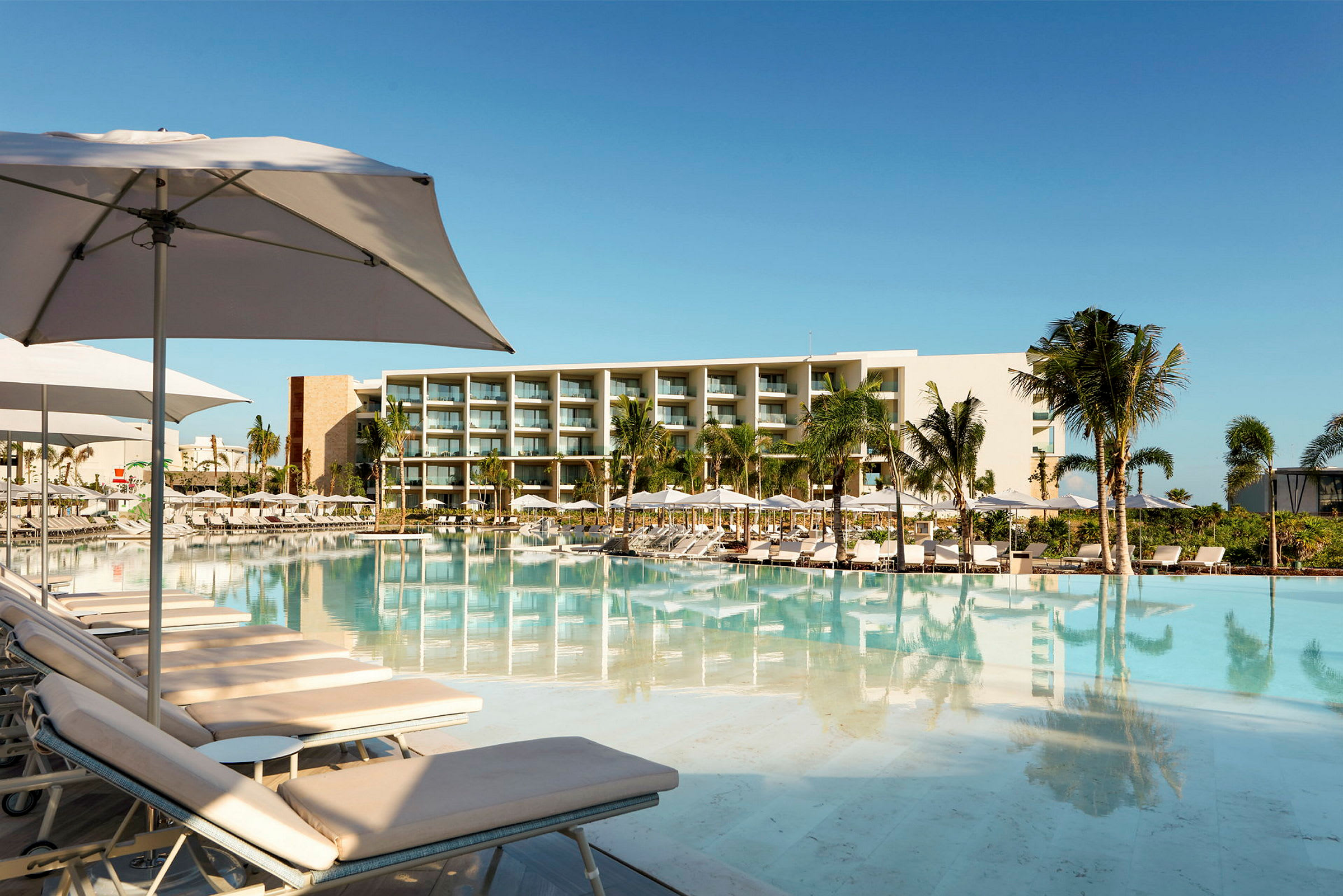 Moderne hotel med stor swimmingpool omgivet af liggestole, parasoller og palmer. (AI-genereret tekst)