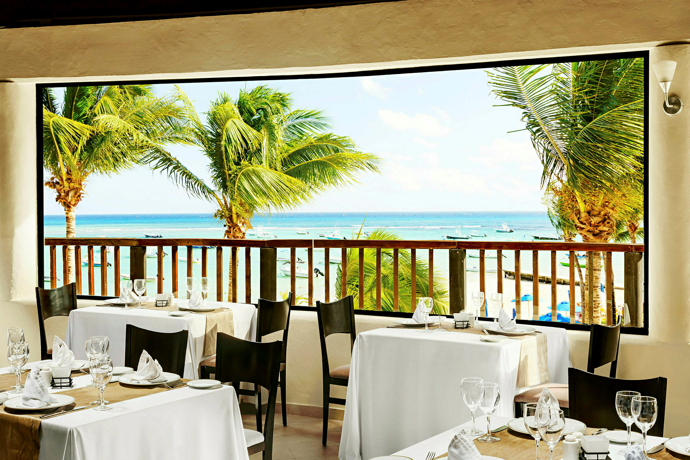 Udsigt fra en restaurant med dækkede borde mod stranden og havet, omgivet af palmer. (AI-genereret tekst)