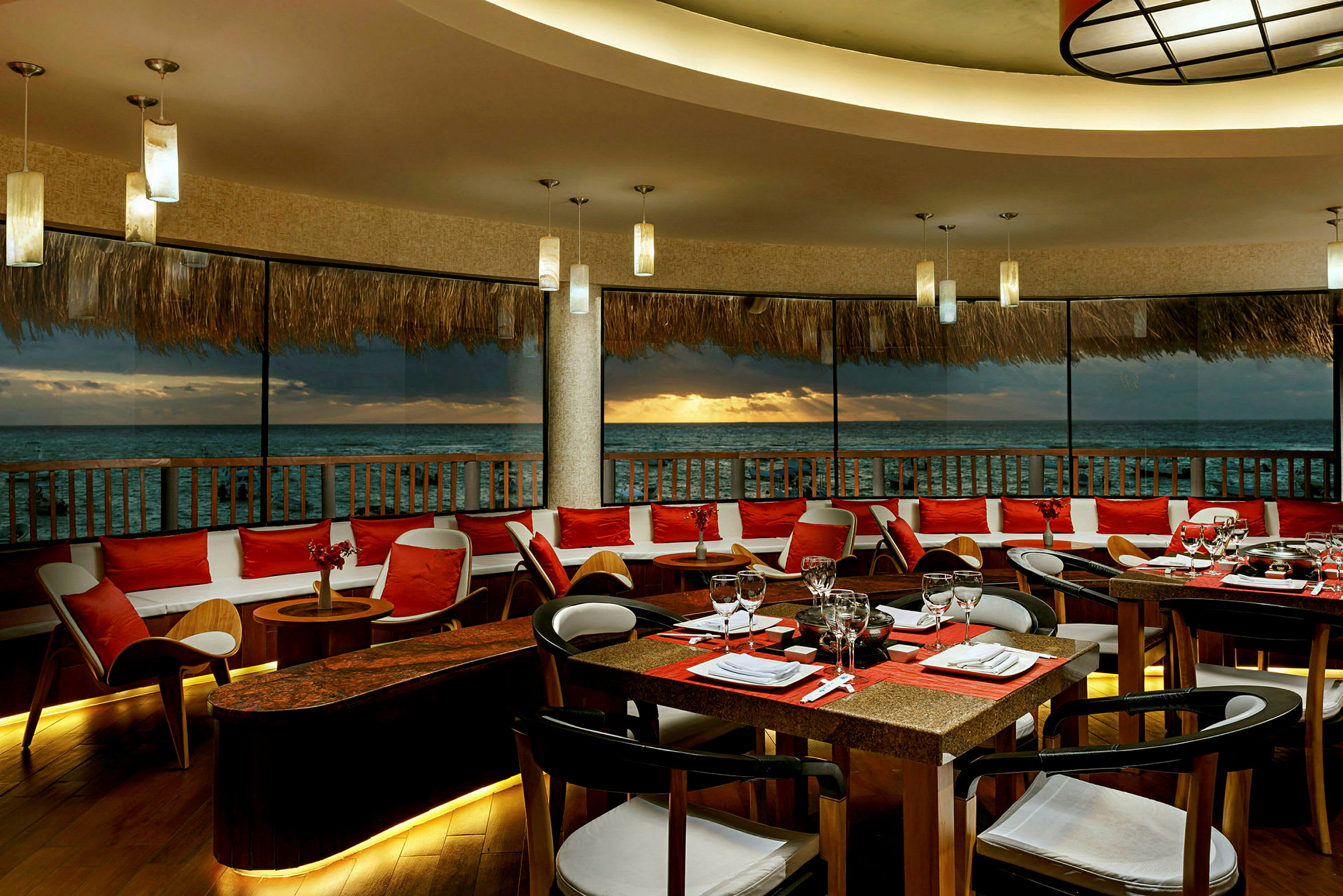 En restaurant med borde og stole placeret ved et stort vindue med udsigt over havet og solnedgangen. (AI-genereret tekst)