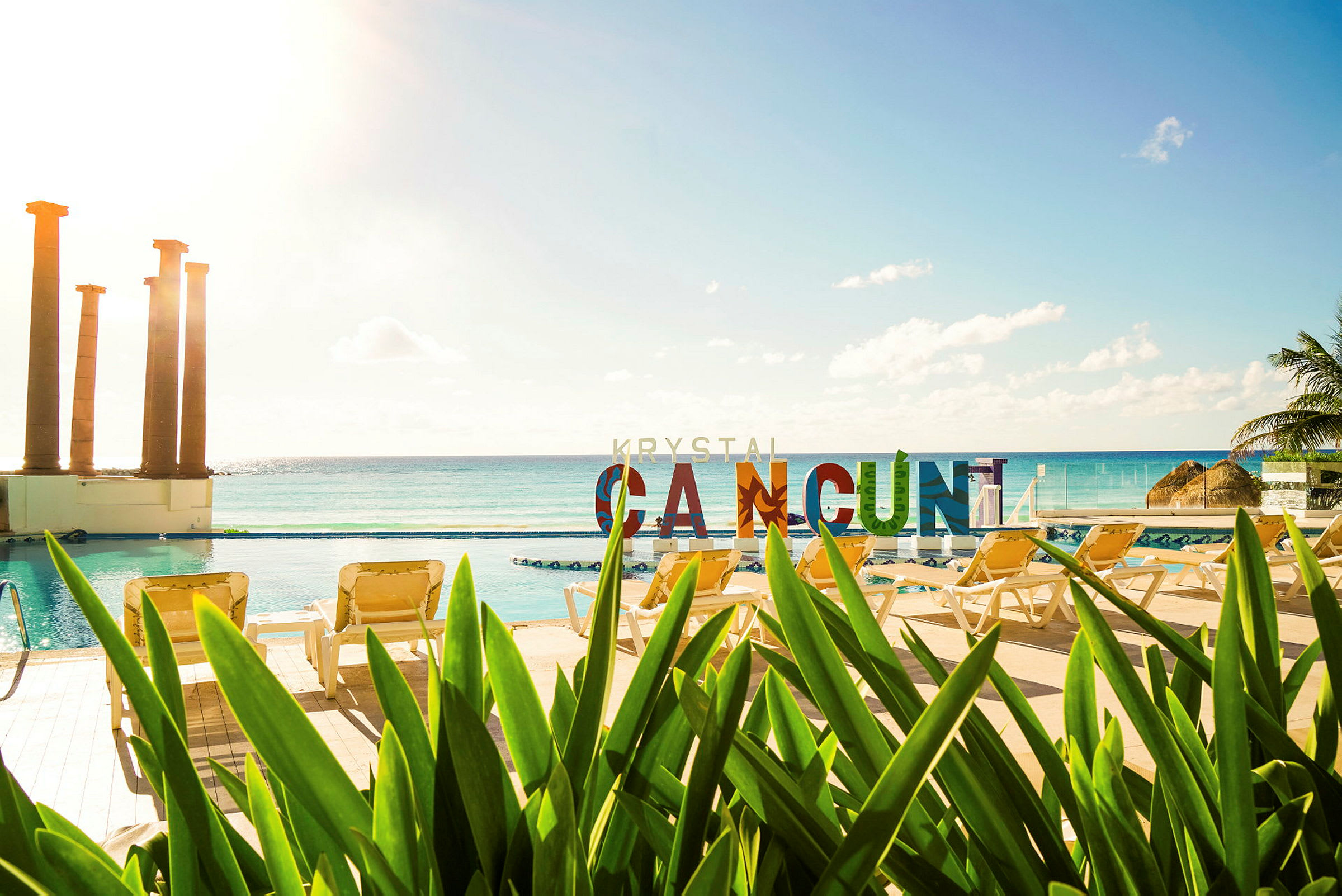 Udsigt over en pool med liggestole og vegetation i forgrunden. I baggrunden ses havet og farverige bogstaver, der staver "CANCUN". (AI-genereret tekst)