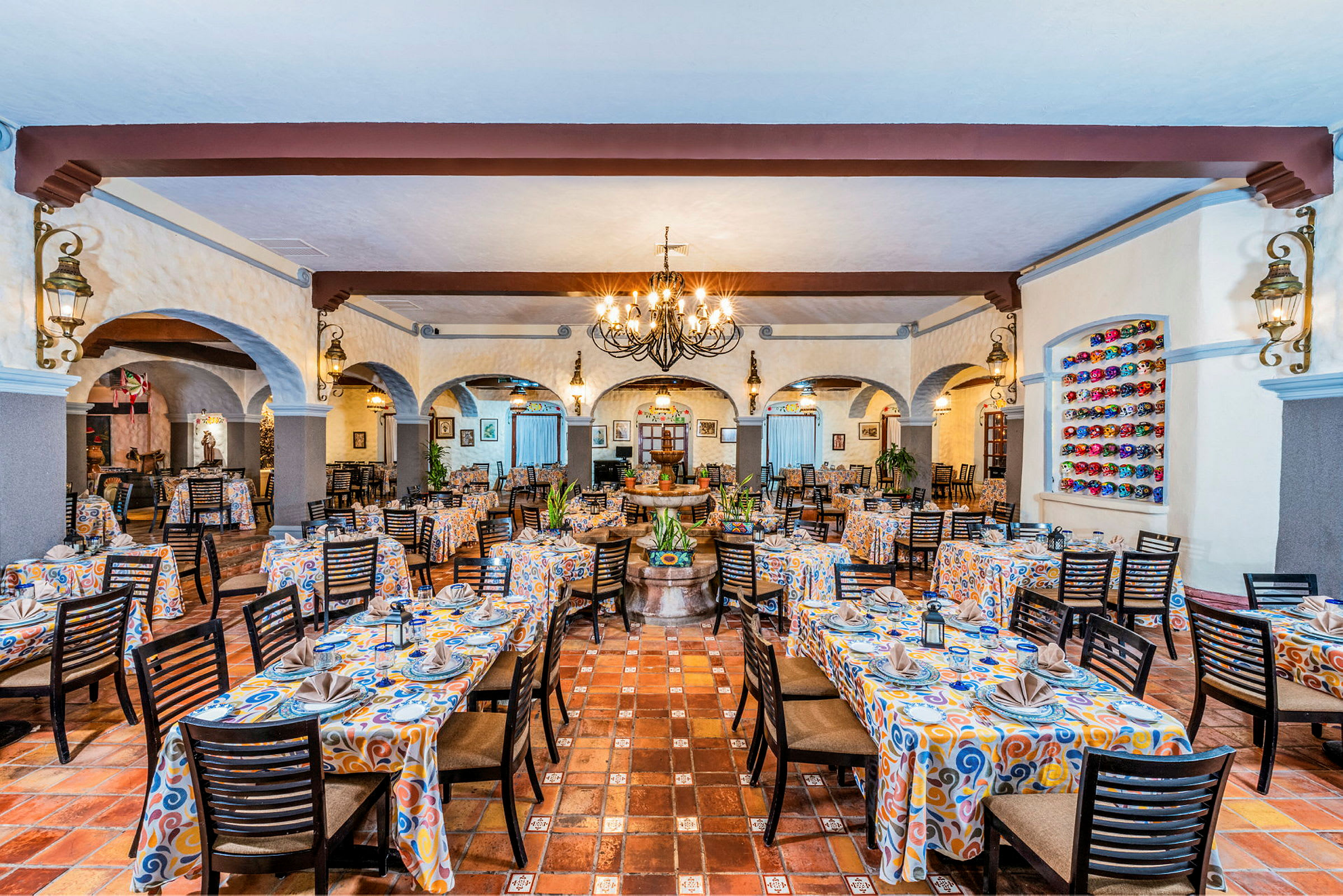 Et rummeligt restaurantområde med borde dækket med mønstrede duge og stole. Der er dekorationer på væggene og en hængende lysekrone i midten. (AI-genereret tekst)