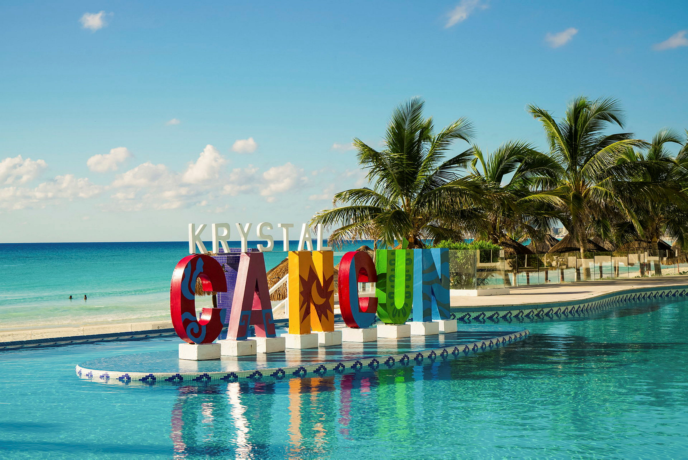 Store farverige bogstaver, der danner teksten 'Cancun' ved en pool. Palmer og hav i baggrunden. (AI-genereret tekst)