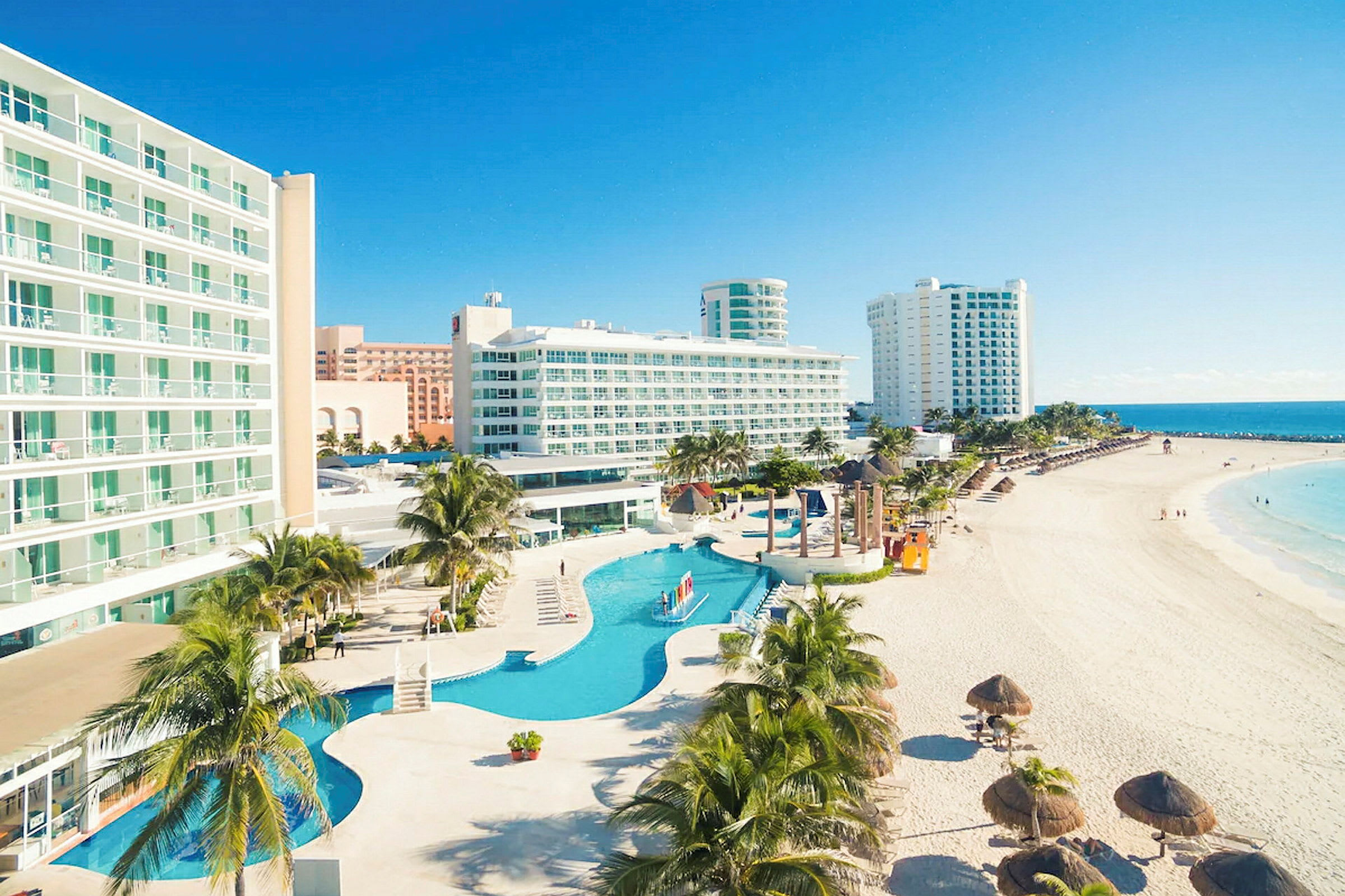 Billede af hotel ved stranden med pool og palmer. (AI-genereret tekst)