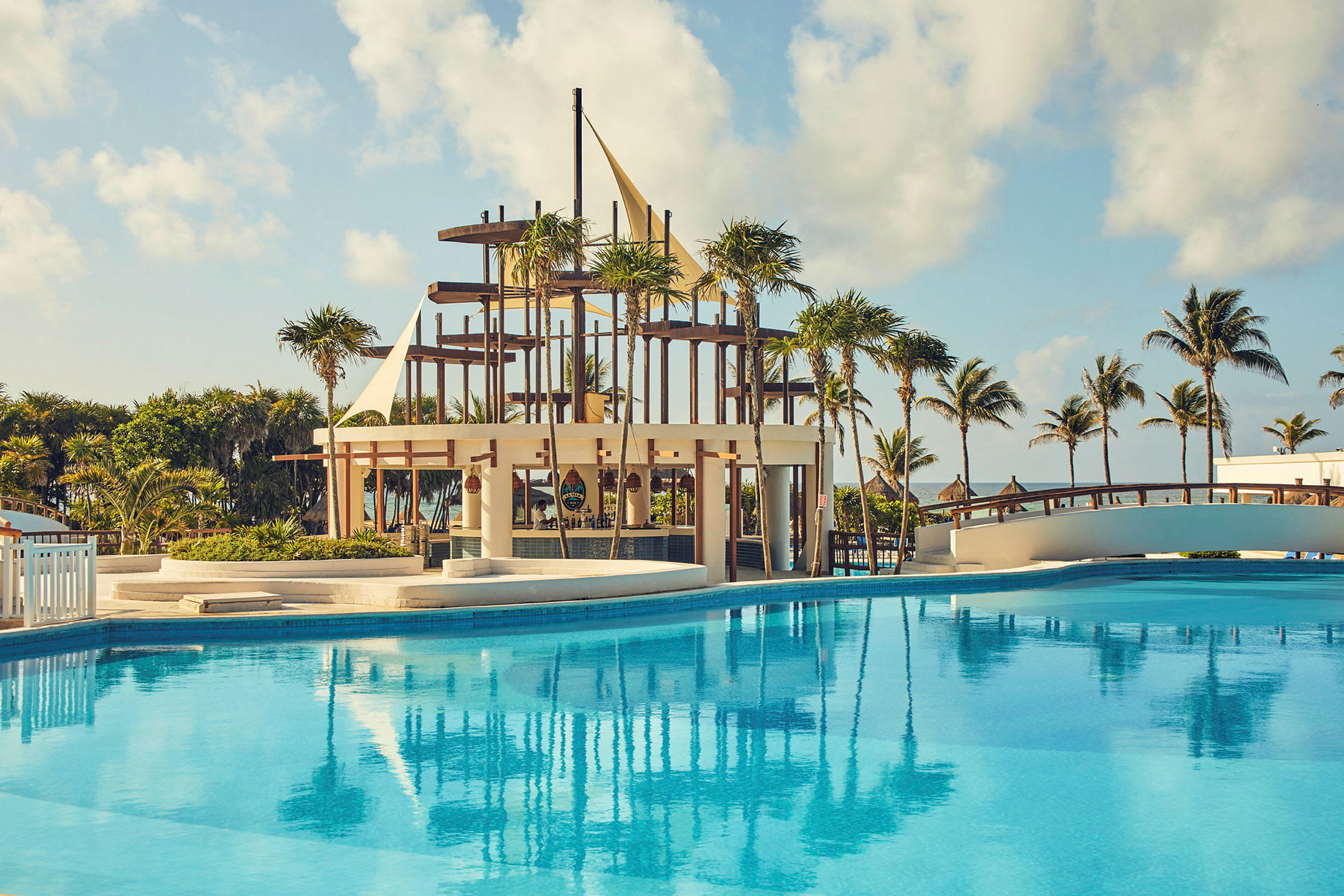 En swimmingpool med klart blåt vand foran en strandbar med trækonstruktioner og sejllignende detaljer. Palmer og en bro ses i baggrunden, samt en klar himmel. (AI-genereret tekst)
