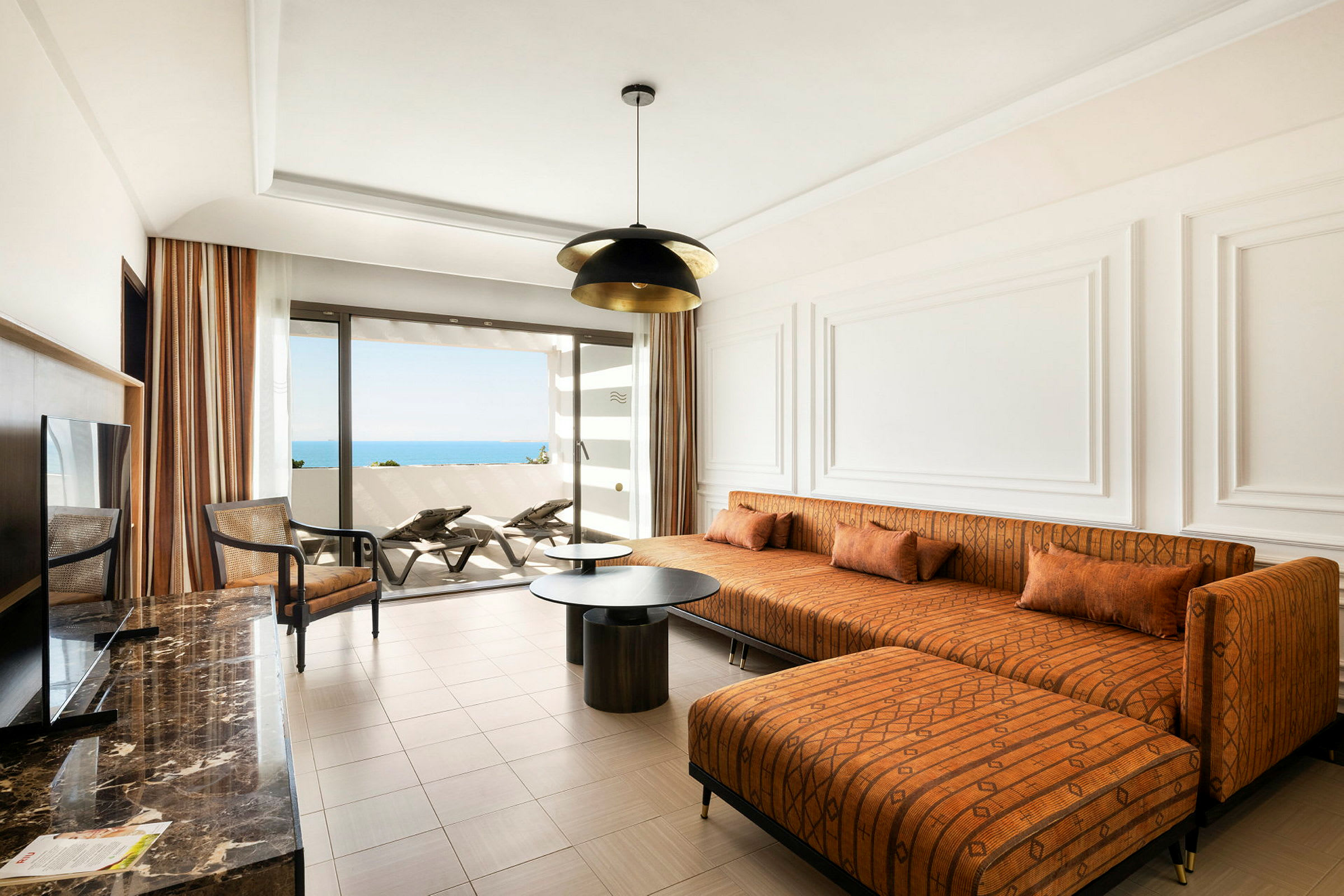 Et moderne hotelværelse med en orange sofa, et sofabord og udsigt til havet fra en balkon. (AI-genereret tekst)