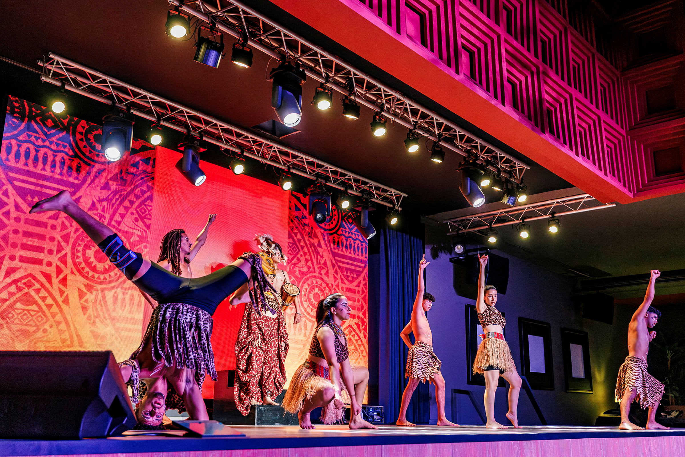 Gruppe af dansere på scenen med lys farverig baggrund og tribal-inspirerede kostumer. (AI-genereret tekst)