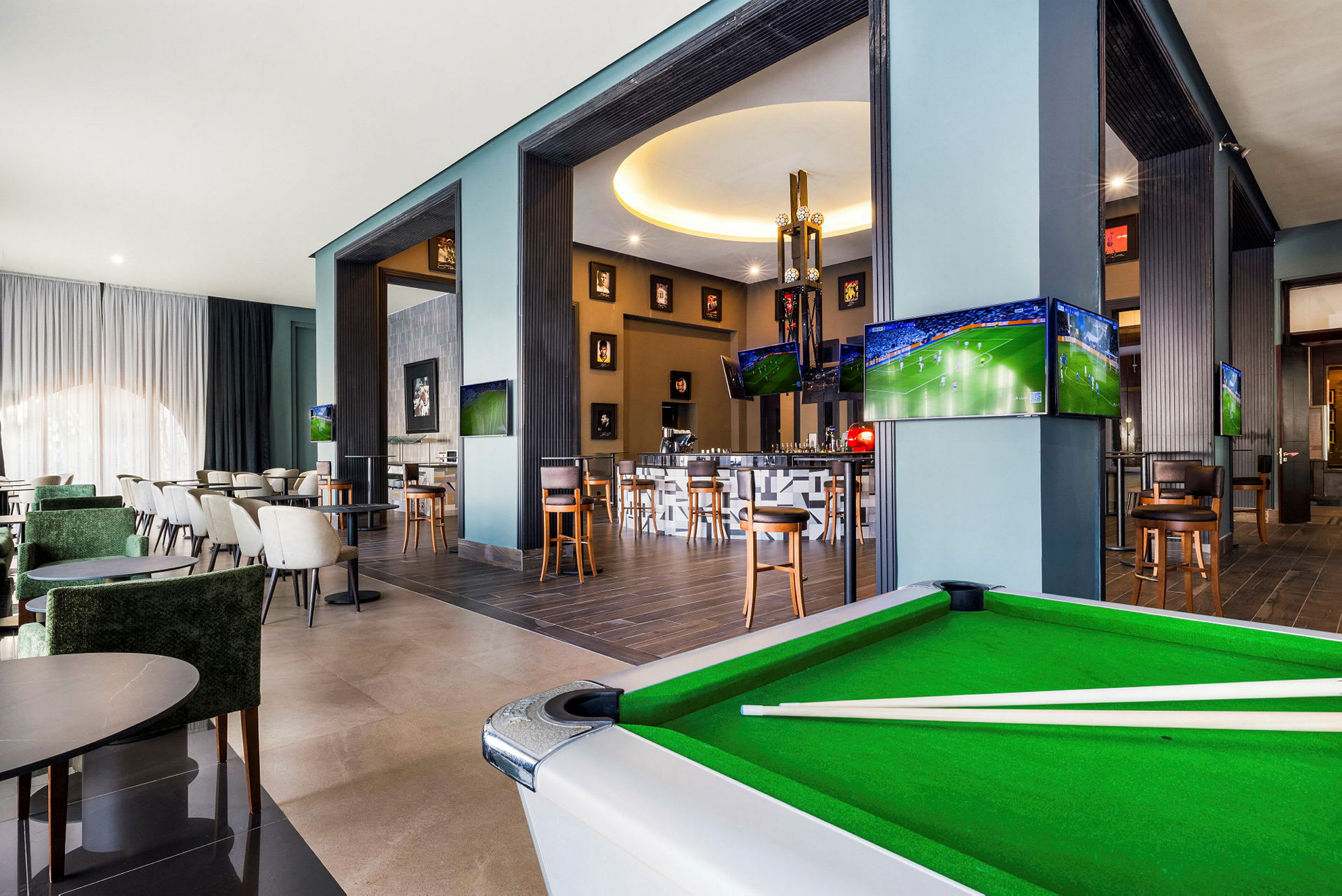 Interiør af en moderne lounge med siddegrupper, et billiardbord og flere TV-skærme, der viser sport. (AI-genereret tekst)