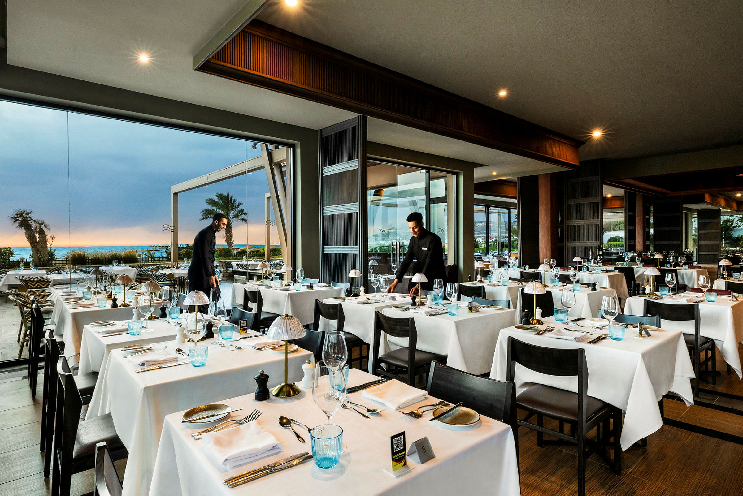 Interiørbillede af en restaurant med dækkede borde og store vinduer, der viser udsigten til havet og palmer. (AI-genereret tekst)