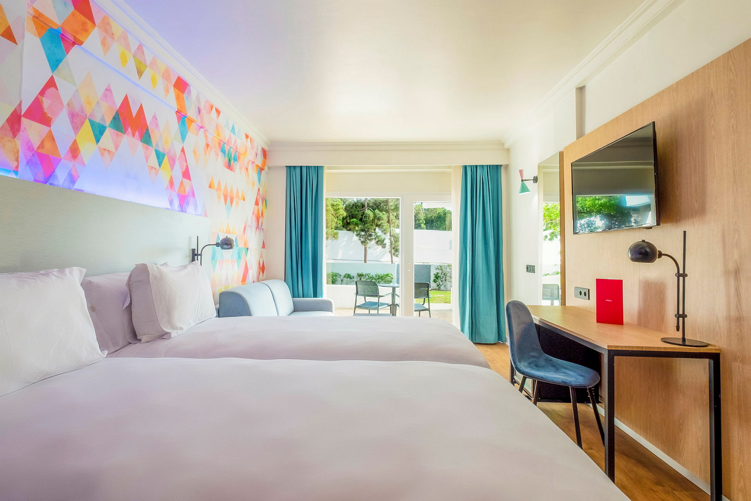 Et hotelværelse med to senge, en farverig vægdekoration, siddeområde, skrivebord og adgang til en terrasse med stole. (AI-genereret tekst)