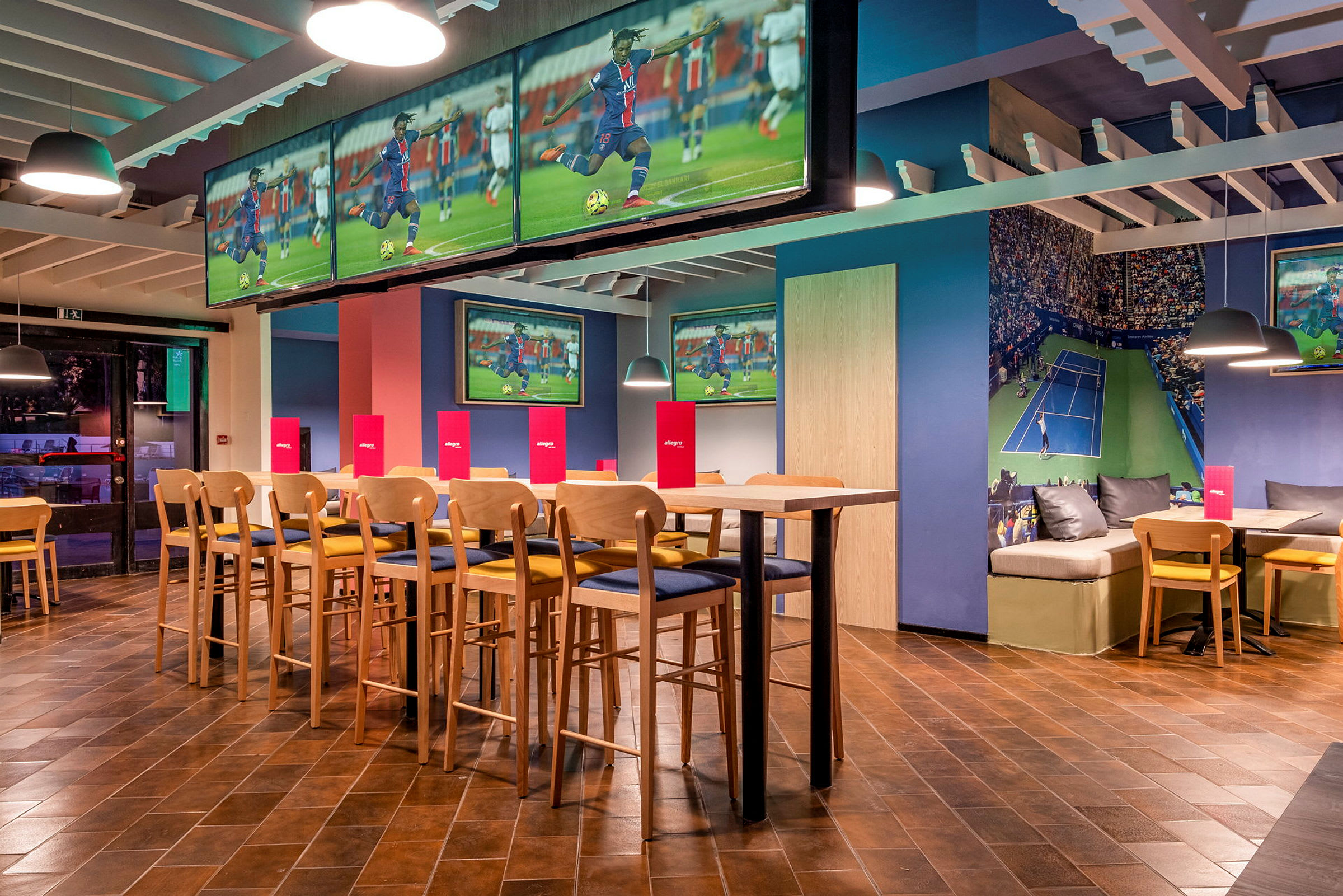 Interiør af en sportsbar med tv-skærme, der viser fodbold og tennis, siddepladser og borde. (AI-genereret tekst)