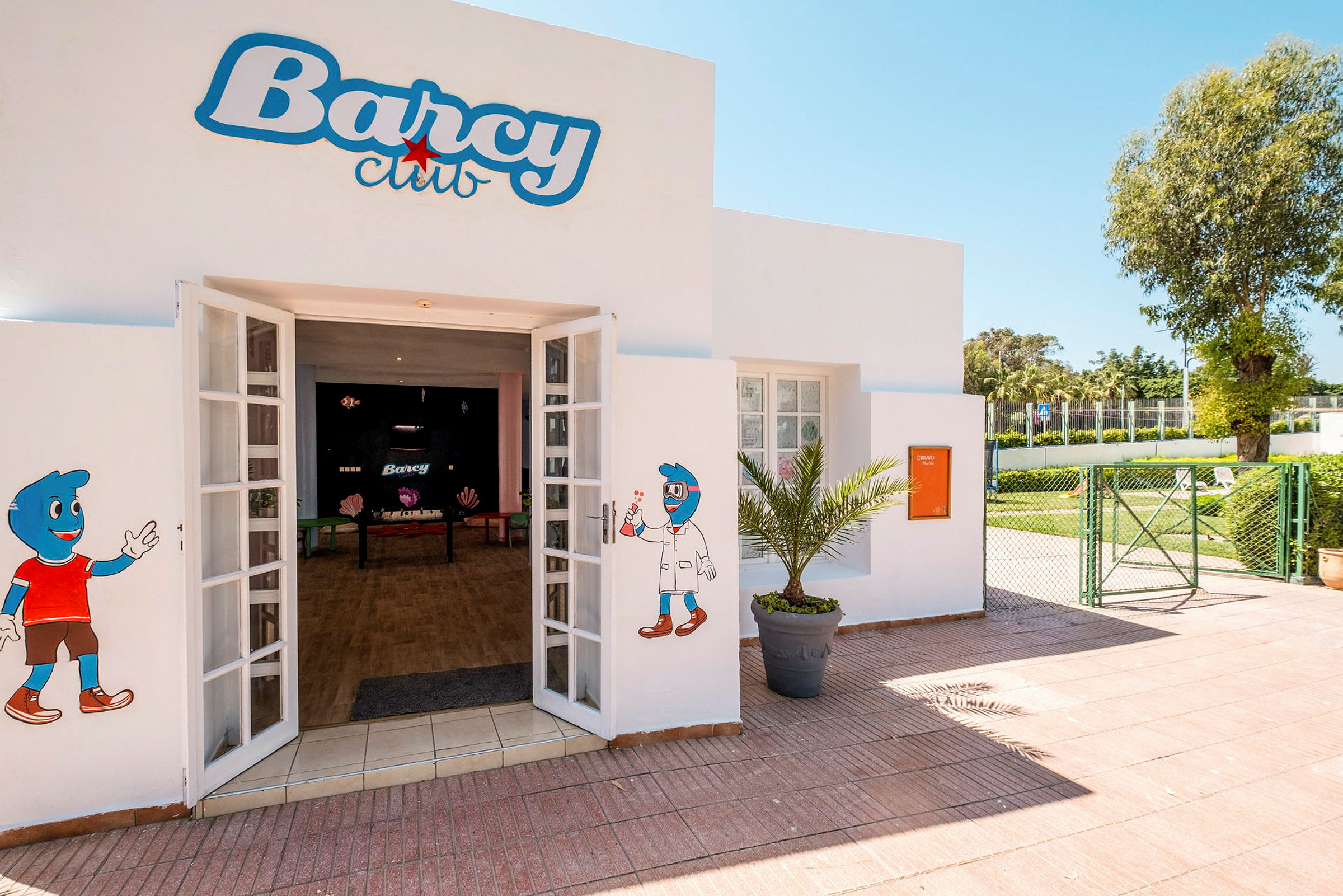 Indgang til Barcy Club med dekorationer på væggene og en gårdsplads med grønne områder. (AI-genereret tekst)