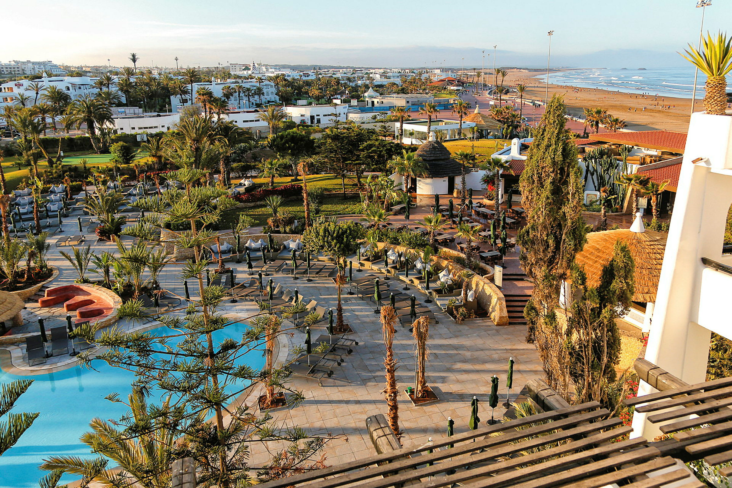 Et hotelområde med pool, liggestole og omkringliggende haver tæt på en strand. (AI-genereret tekst)