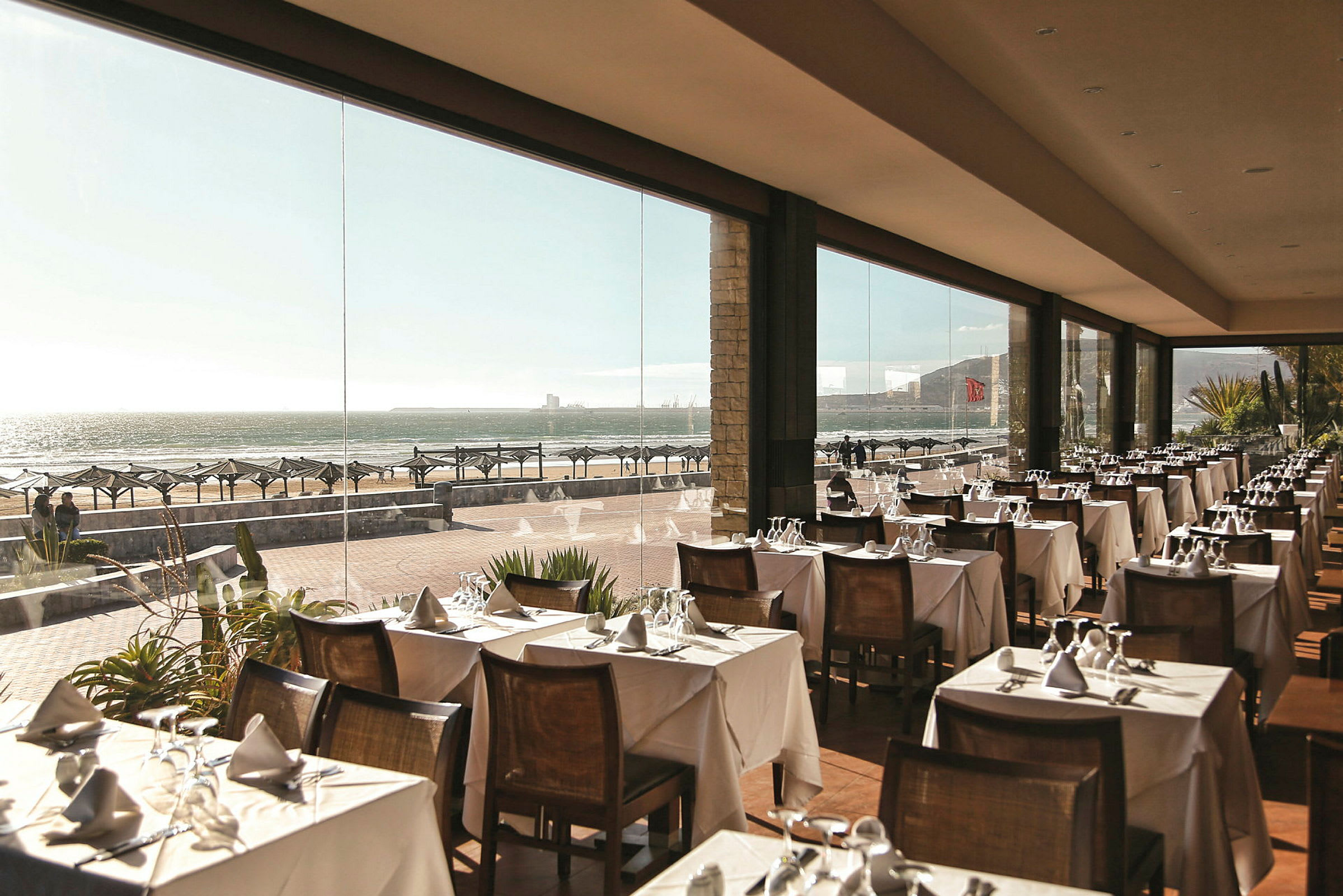 Restaurant med dækket bord og glasvægge med udsigt over stranden. (AI-genereret tekst)