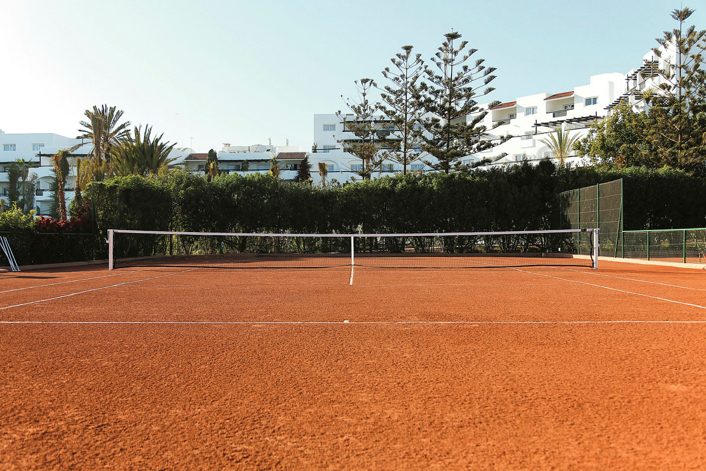Billede af en tennisbane med grusunderlag, omgivet af træer og bygninger i baggrunden. (AI-genereret tekst)