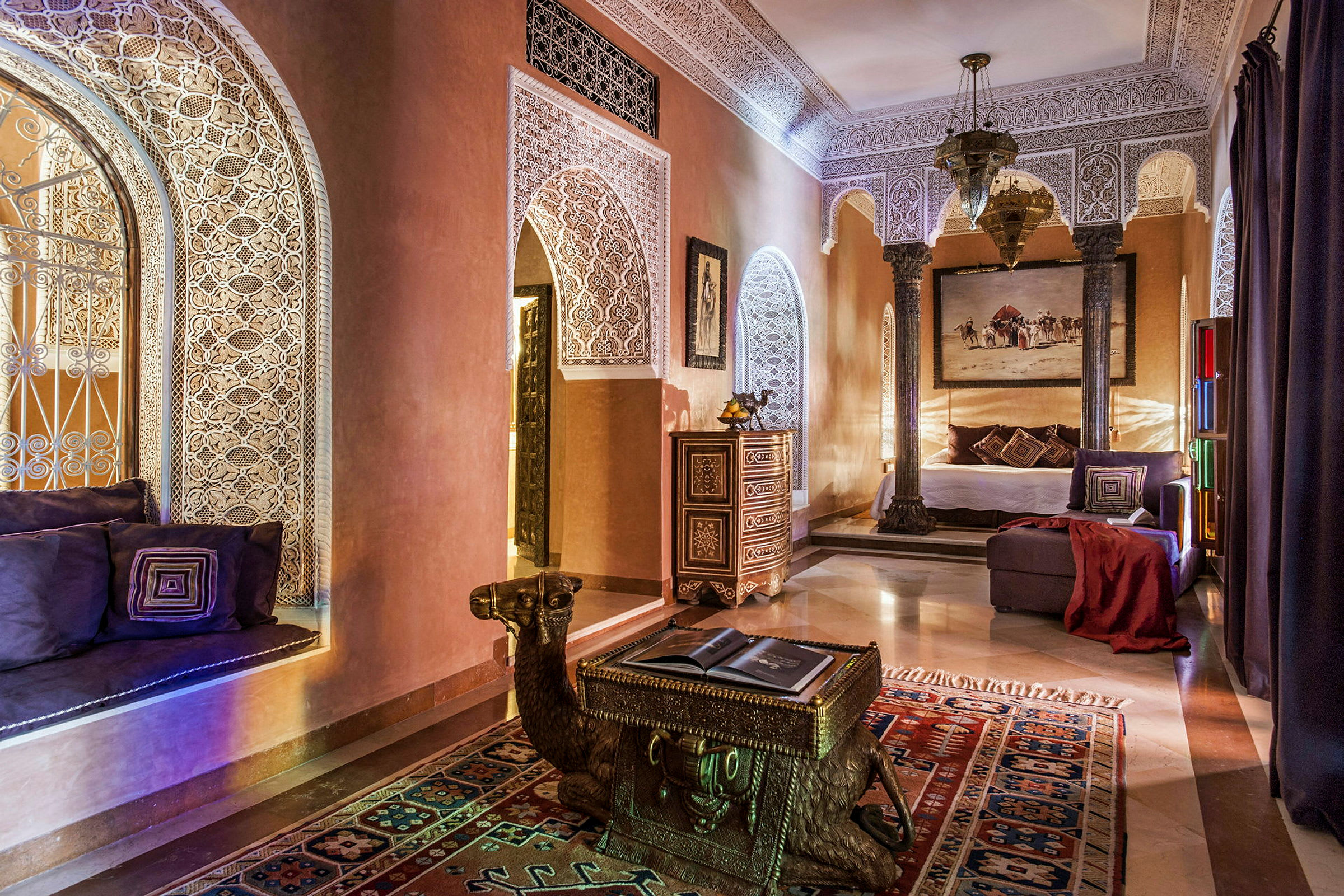 Et elegant indrettet rum med dekorative gipsmønstre, arkitektoniske buer, orientalske møbler og varm belysning. Et bord formet som en kamel står på et farverigt tæppe. (AI-genereret tekst)